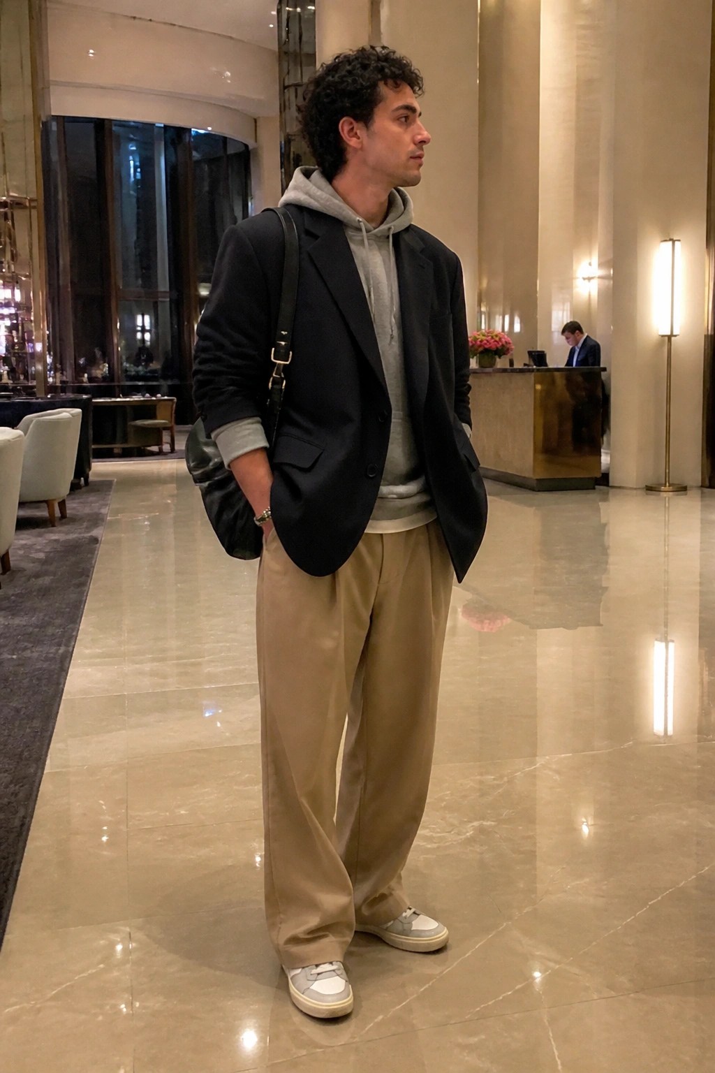 Curly-haired man in black blazer over grey hoodie with beige pants in lobby.