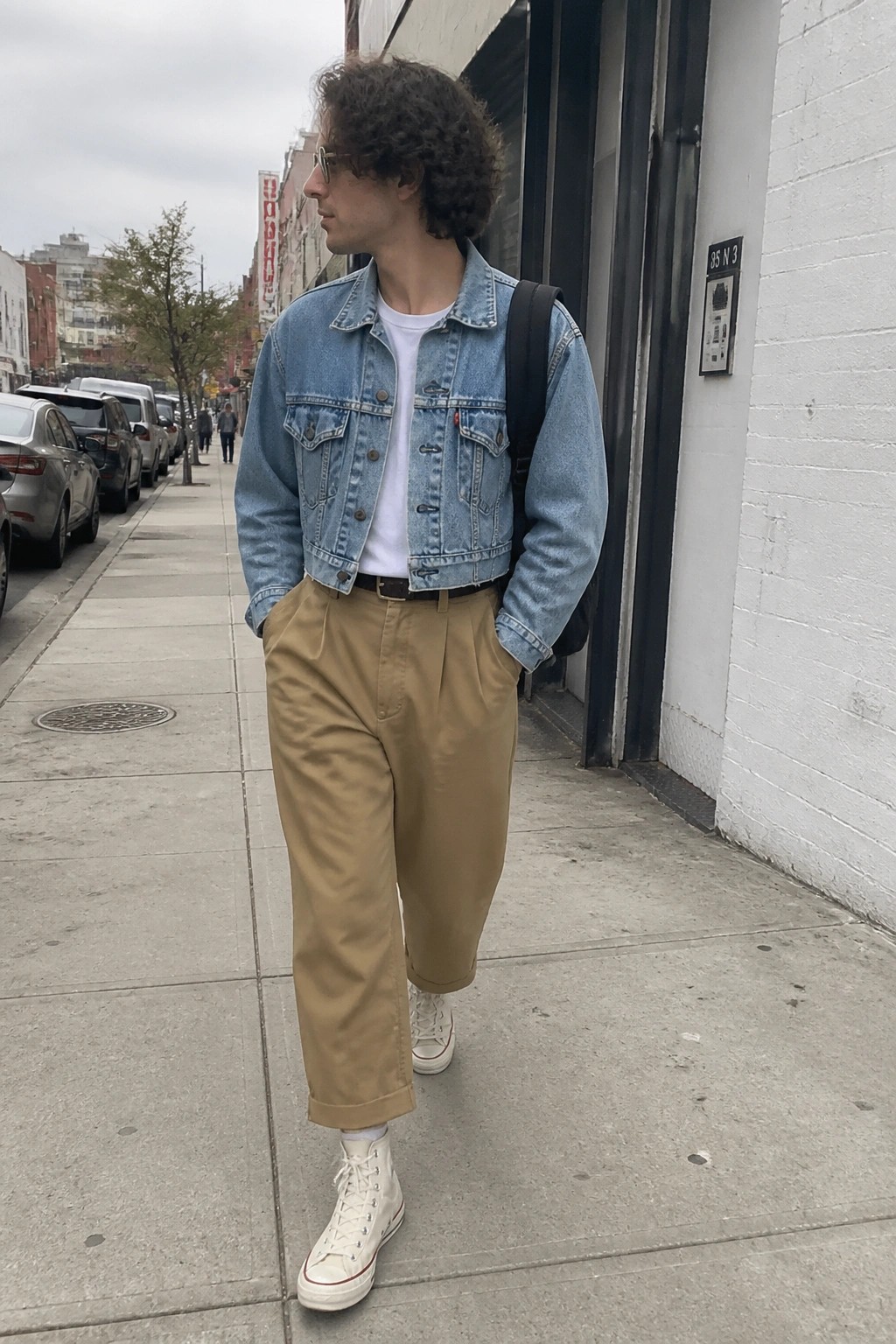 Curly-haired man in denim jacket and khaki pants stands on city sidewalk with backpack