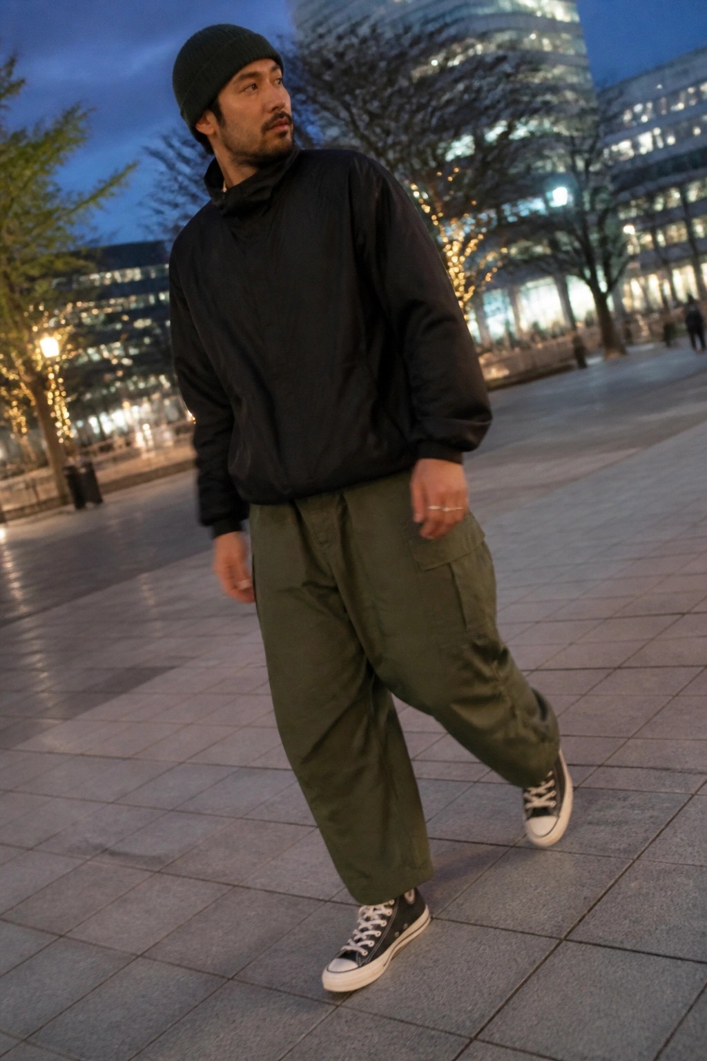 Man in black beanie and jacket with green cargo pants on nighttime city plaza.