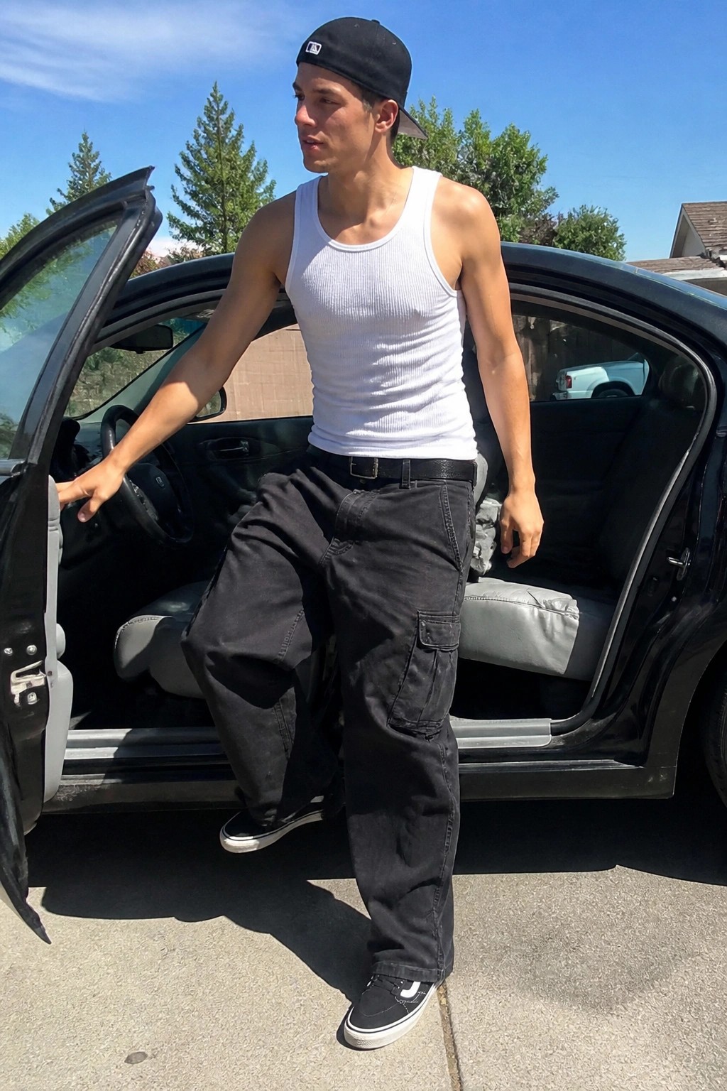 Man in white tank top, black cap and cargo pants by open black car door
