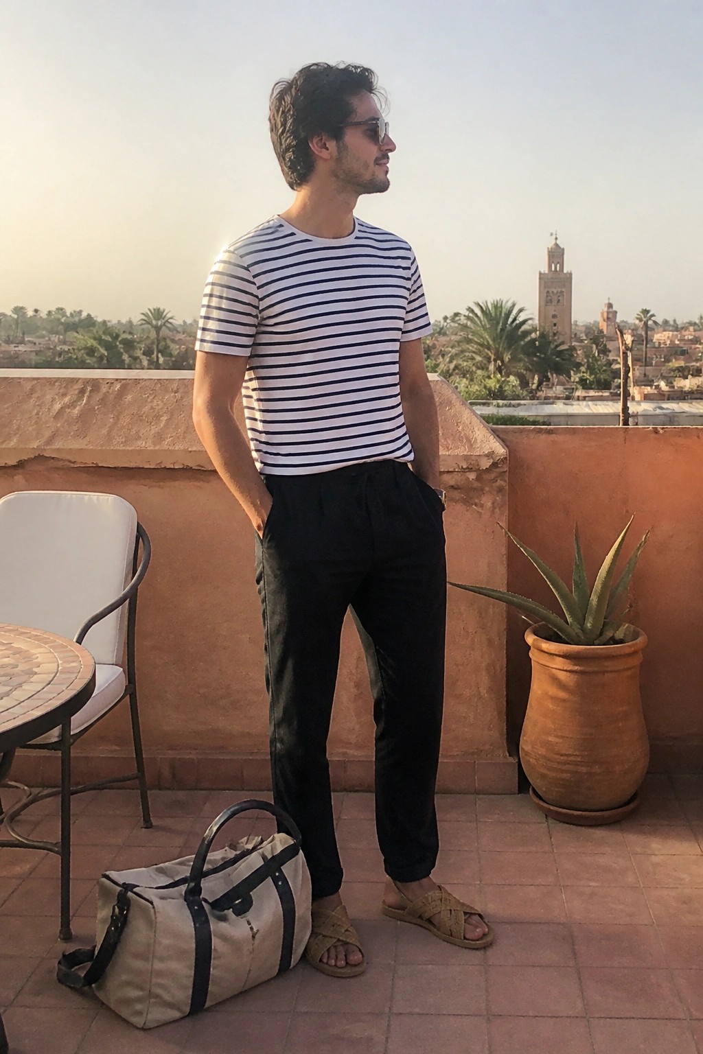 Man in striped shirt and black pants on rooftop with duffel bag and plant