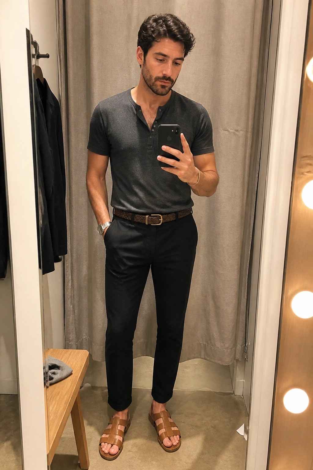 Man in gray henley shirt and black pants taking mirror selfie in fitting room