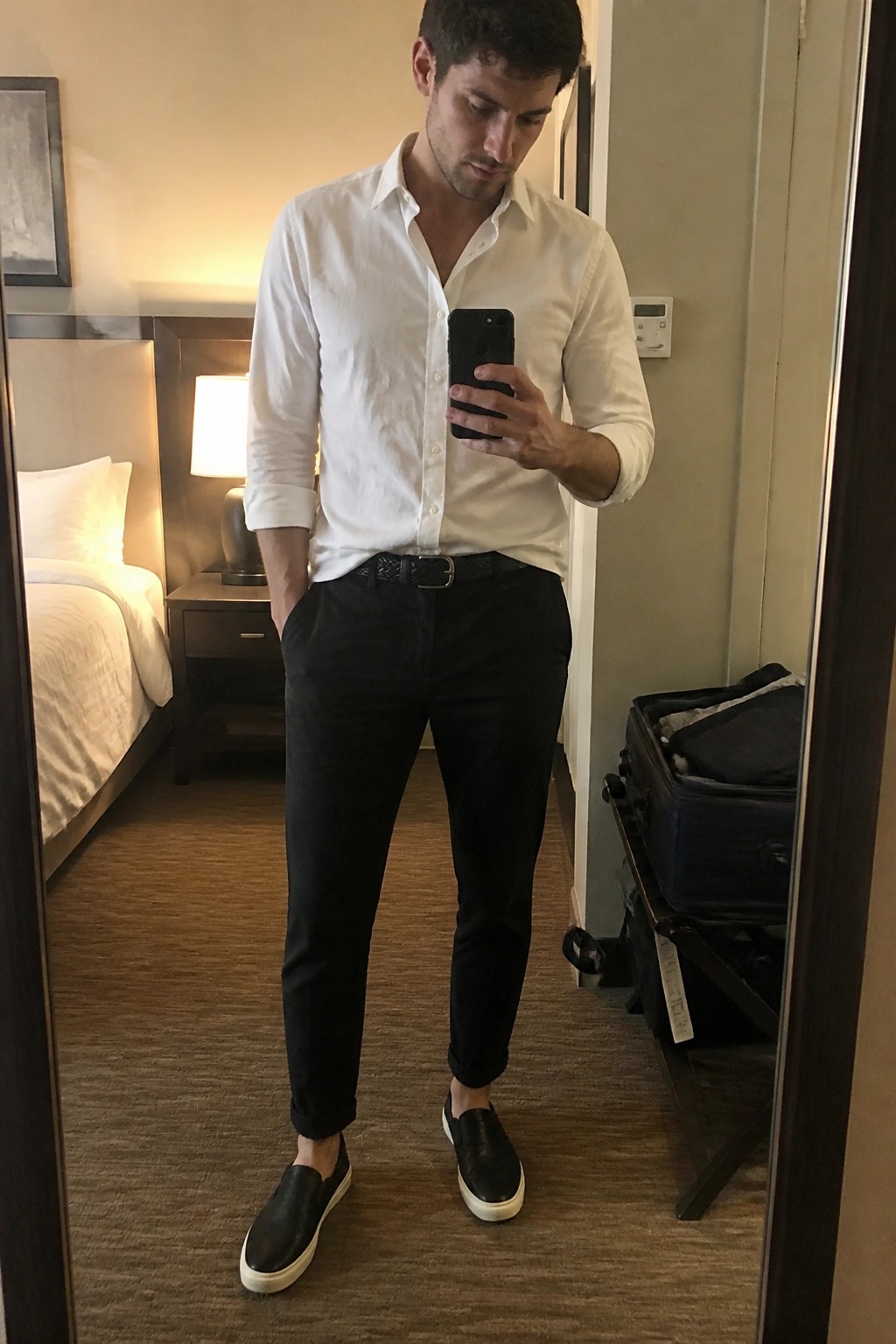 Man in white shirt and black pants taking mirror selfie in hotel room