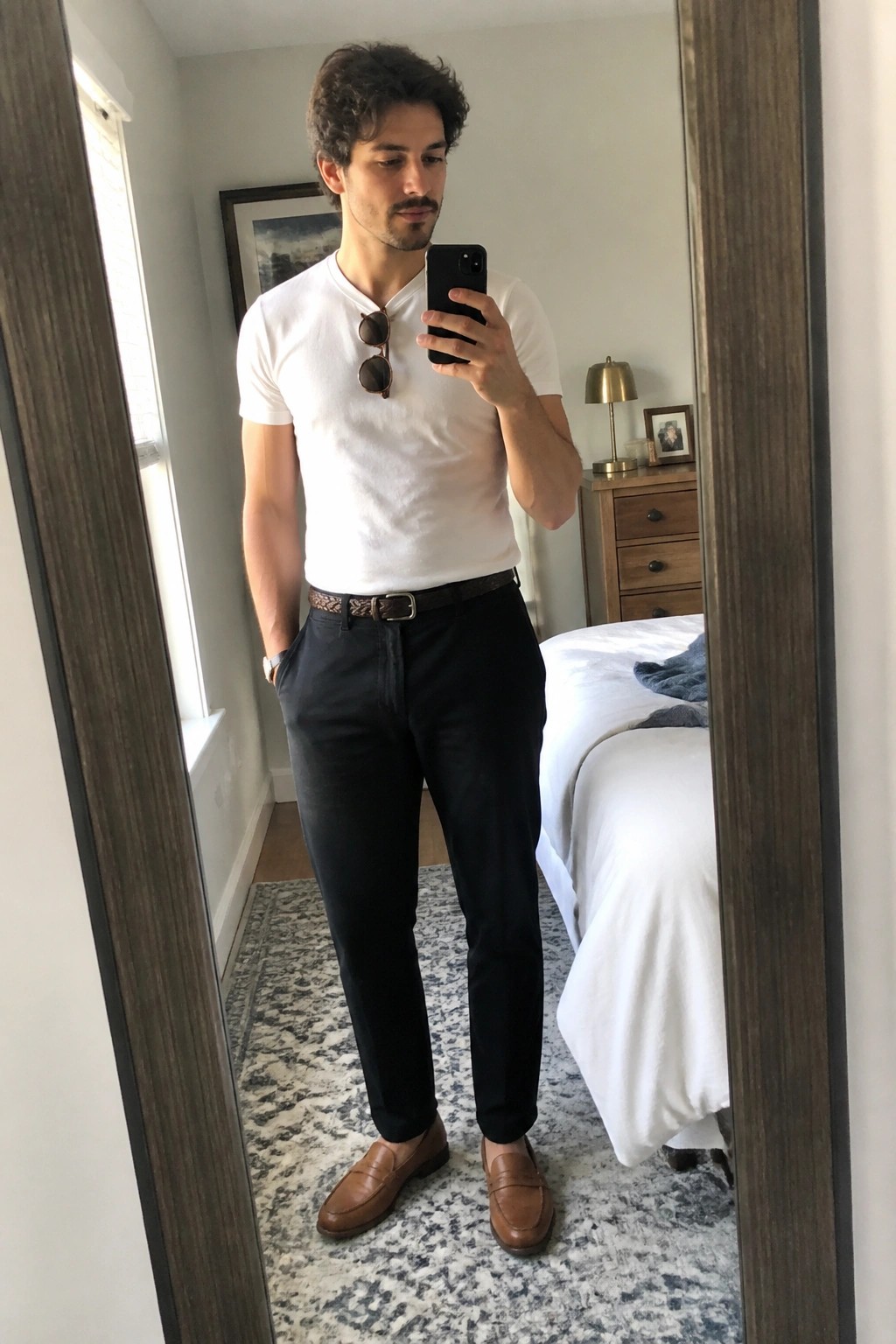 Man in white t-shirt and black pants taking mirror selfie in bedroom