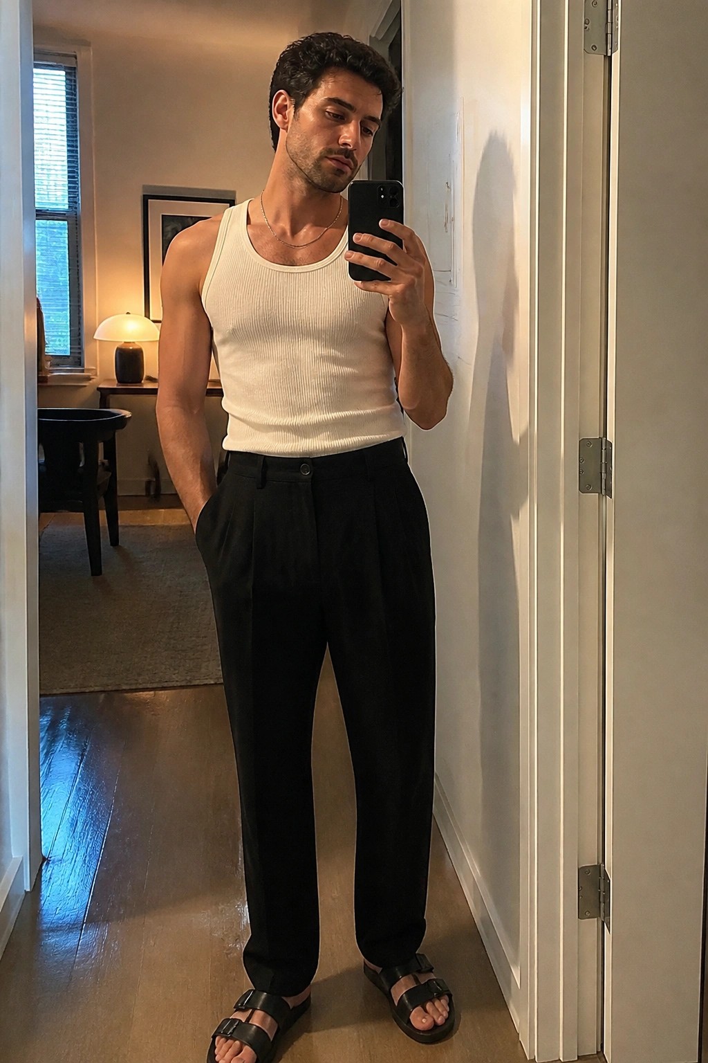 Man in white tank top and black pleated pants taking mirror selfie indoors.