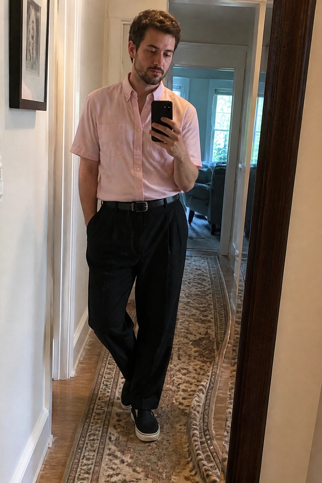Man in pink short-sleeve shirt and black pants taking mirror selfie in hallway