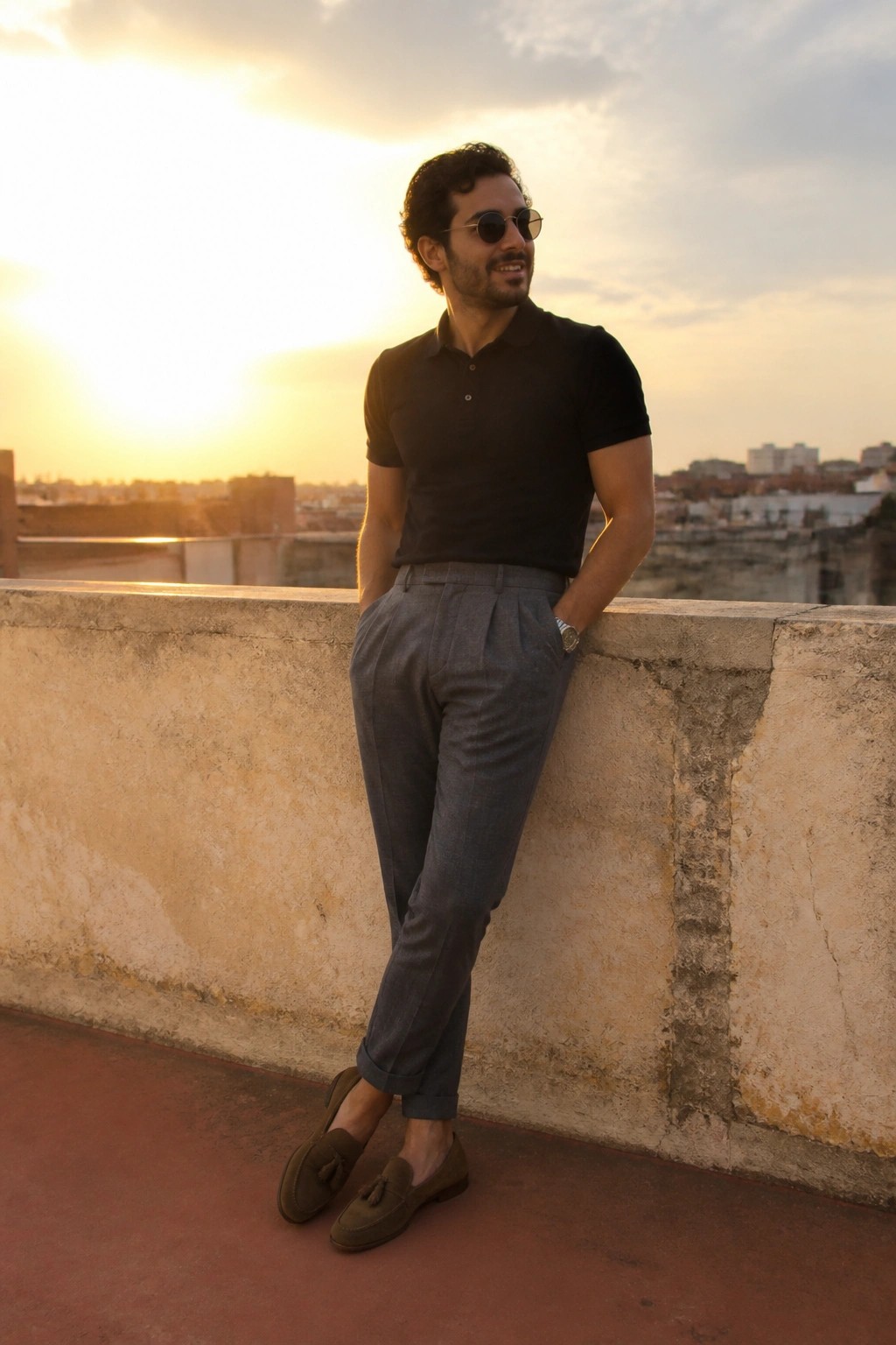 Man in black polo shirt and gray trousers leaning against wall at sunset.