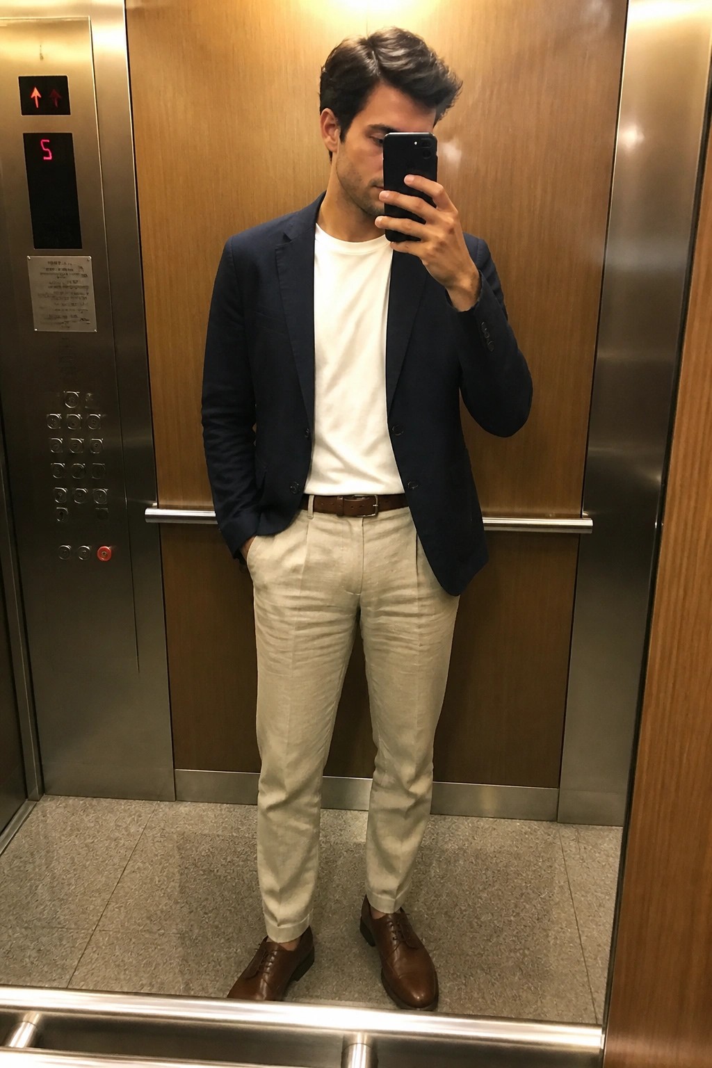 Man in navy blazer and beige pants taking mirror selfie in elevator