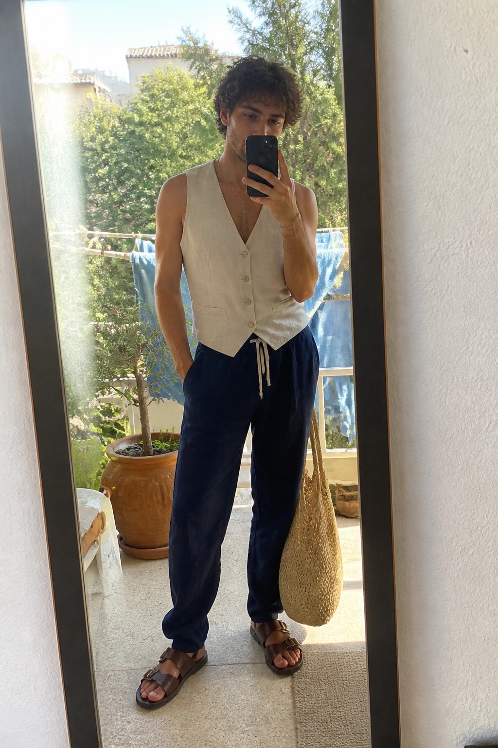 Man in beige vest and blue pants taking mirror selfie on balcony with straw bag