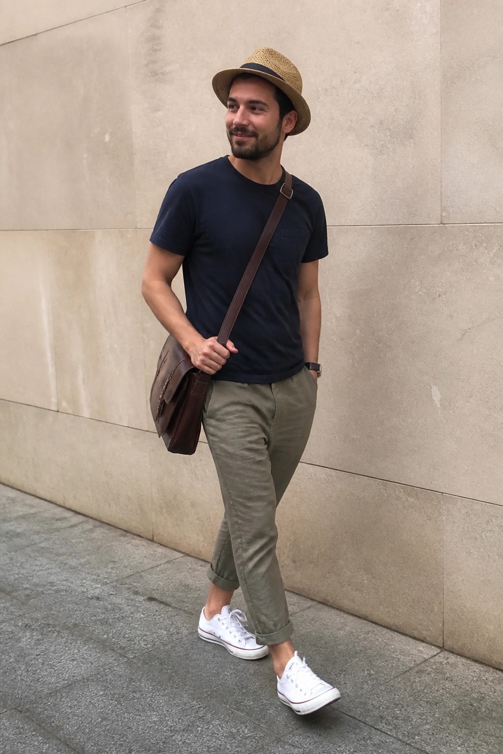 Man in straw hat, navy tee, olive pants with brown leather messenger bag on stone wall