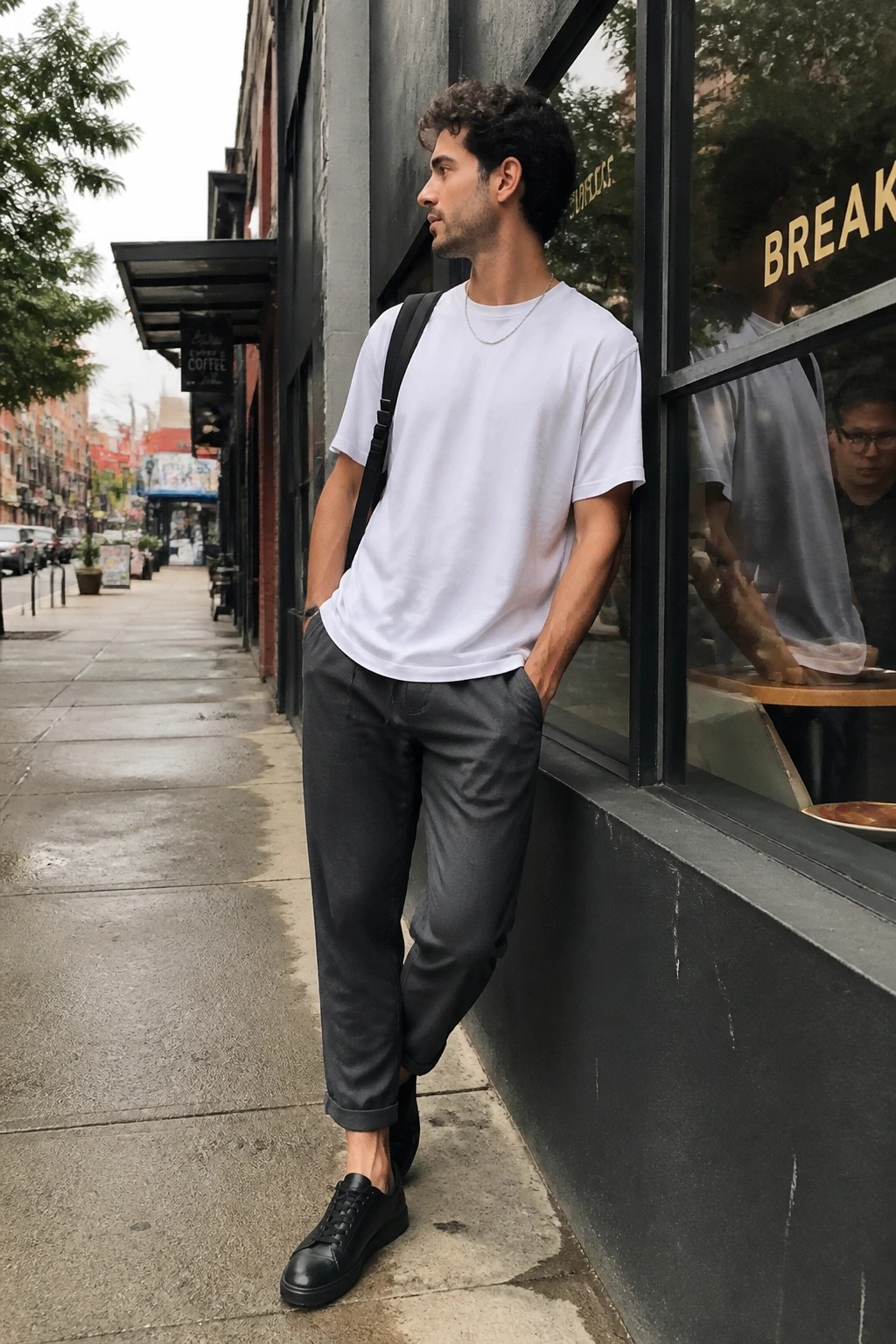 Man in white t-shirt and gray pants leaning against urban cafe window.