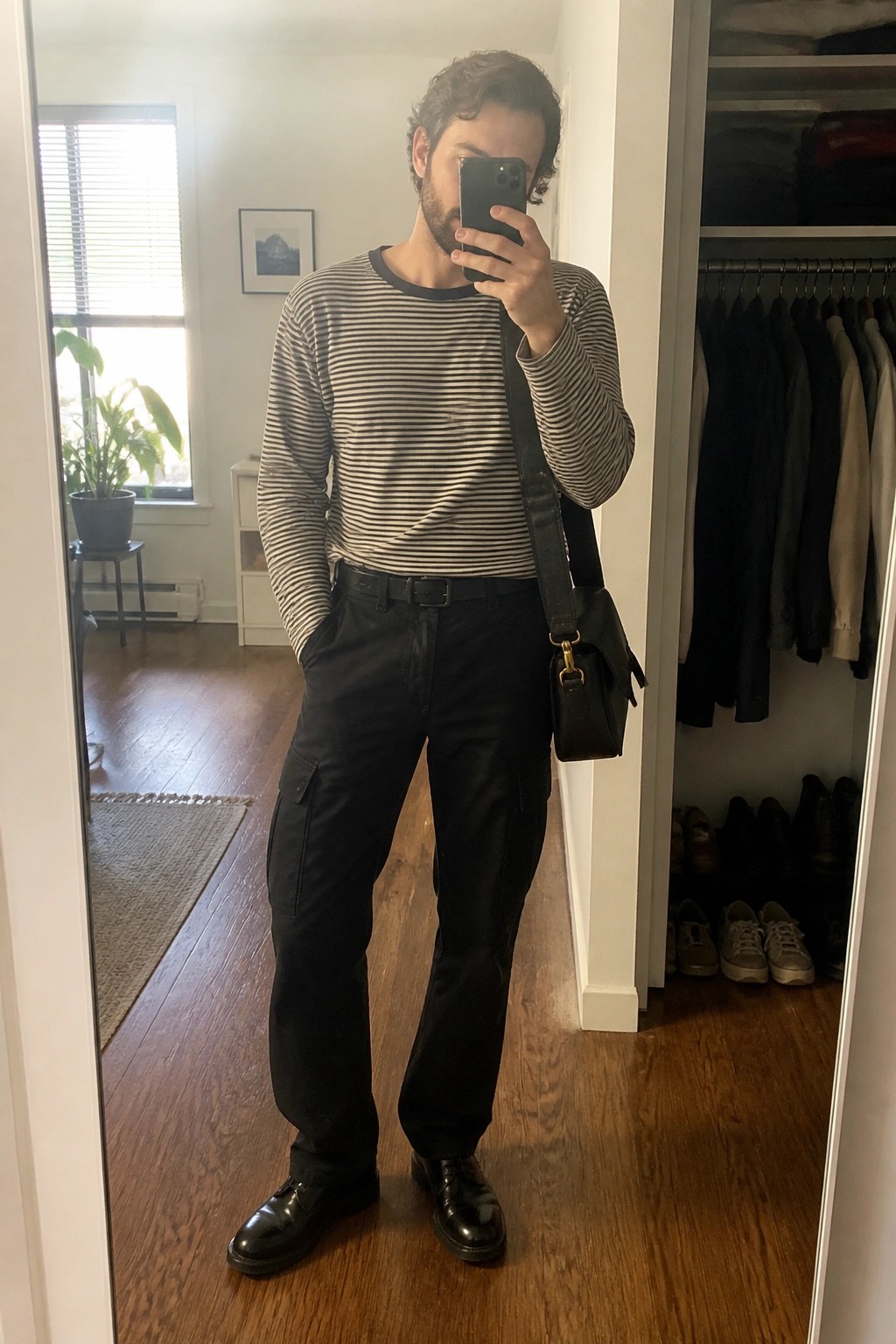 Man in striped shirt and black cargo pants taking mirror selfie with bag.