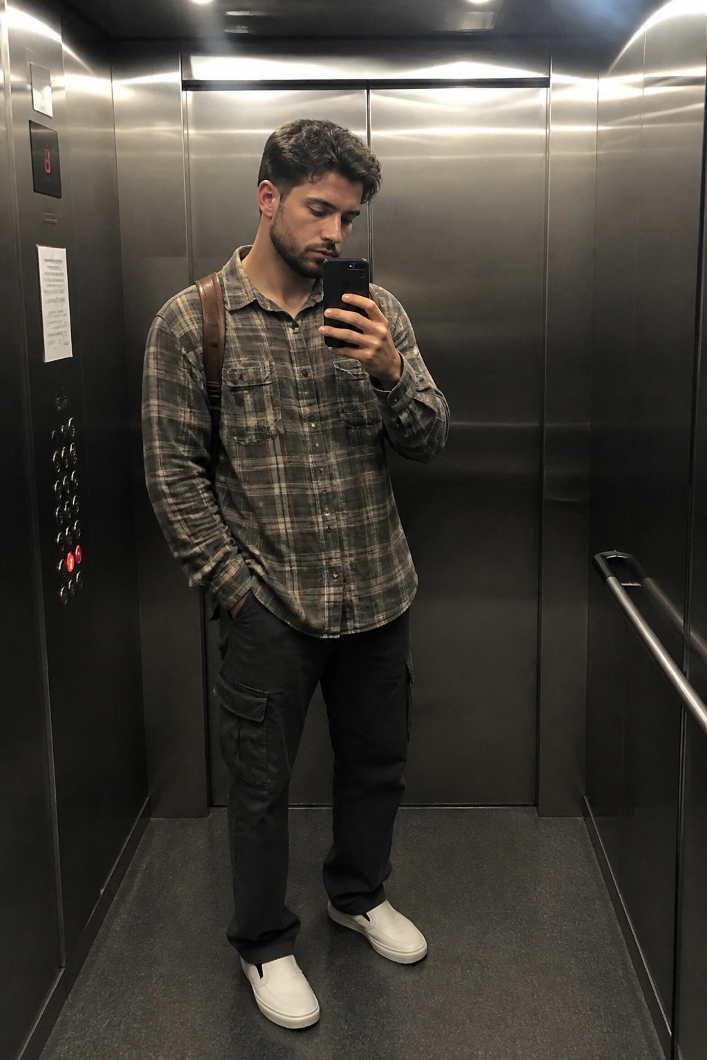 Man in plaid shirt and cargo pants taking mirror selfie in elevator