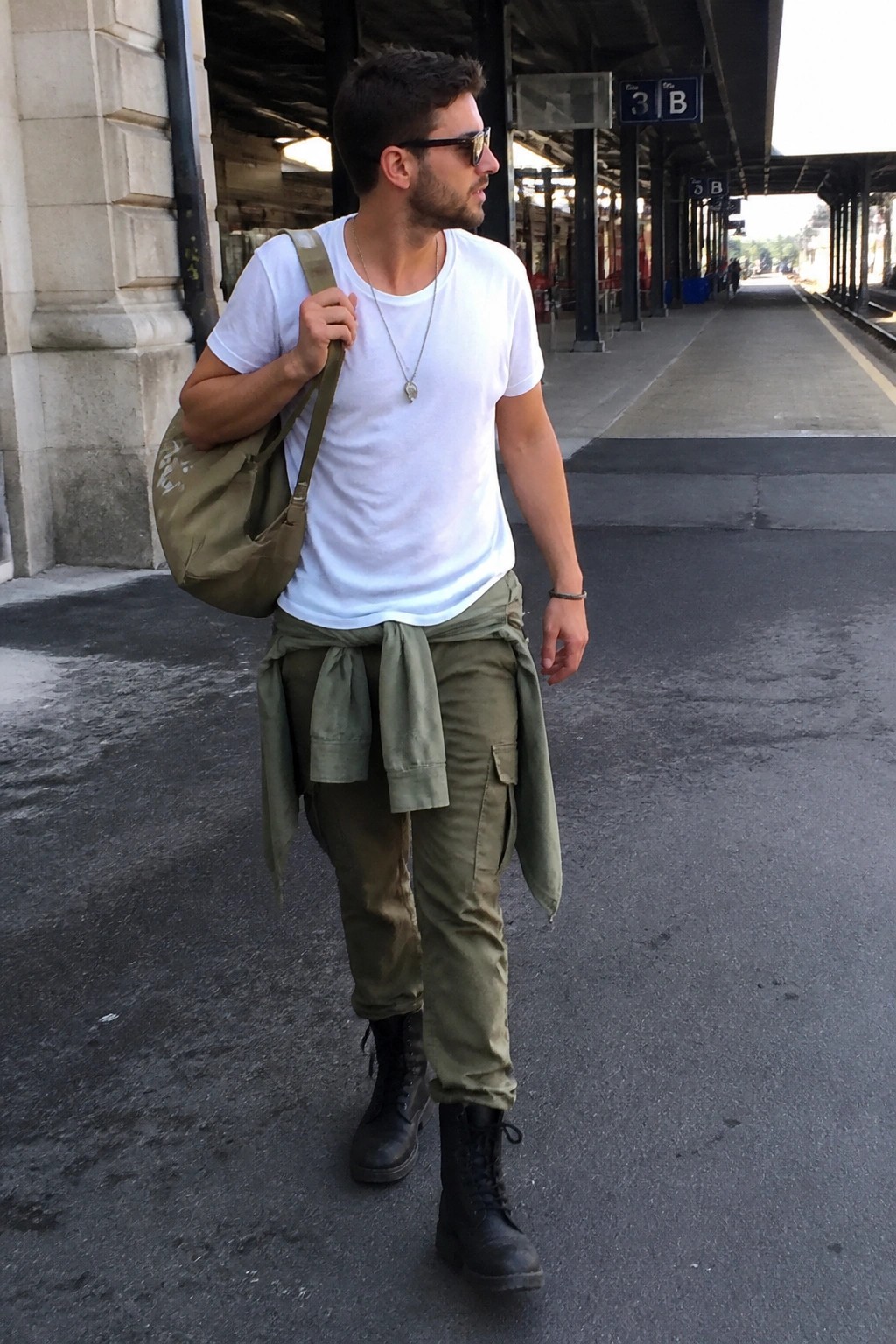 Man in white t-shirt and olive cargo pants with bag on train platform.