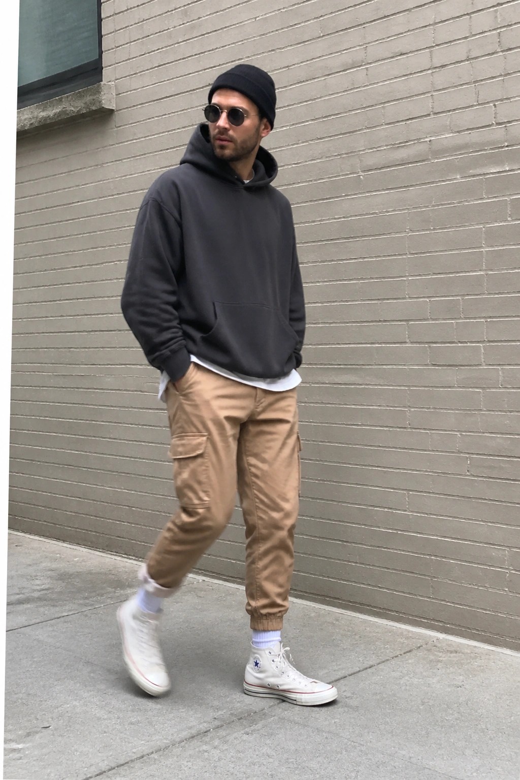 Man in black hoodie and beanie with tan cargo pants and white high-top sneakers by brick wall.