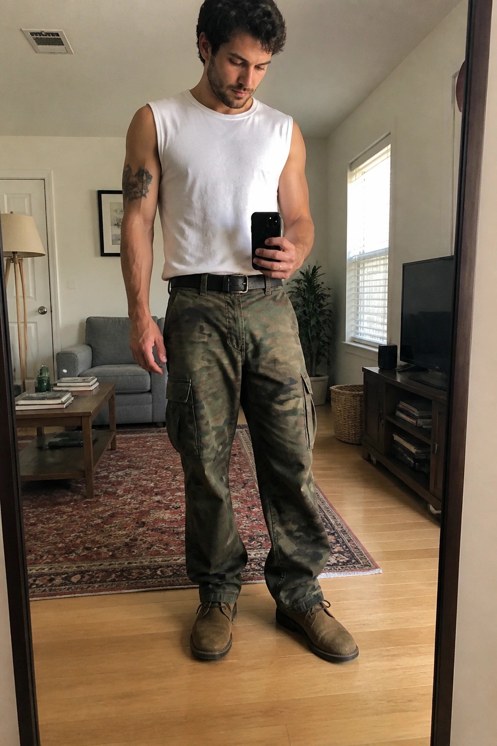Muscular man in white tank top and camouflage cargo pants holding phone in living room