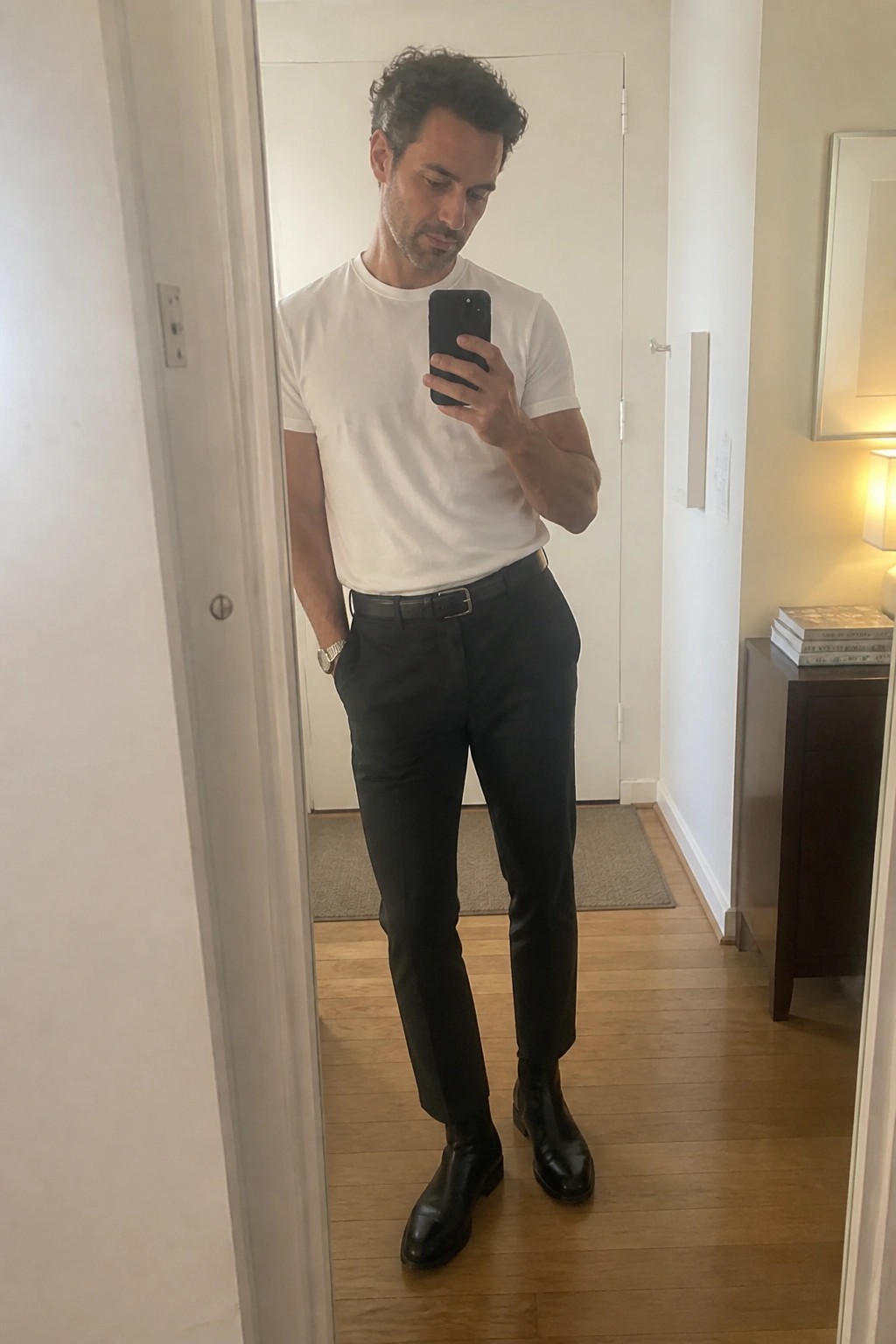 Man wearing slim black pants, white short-sleeve crewneck tee, black leather belt, and black Chelsea boots while taking a mirror selfie.