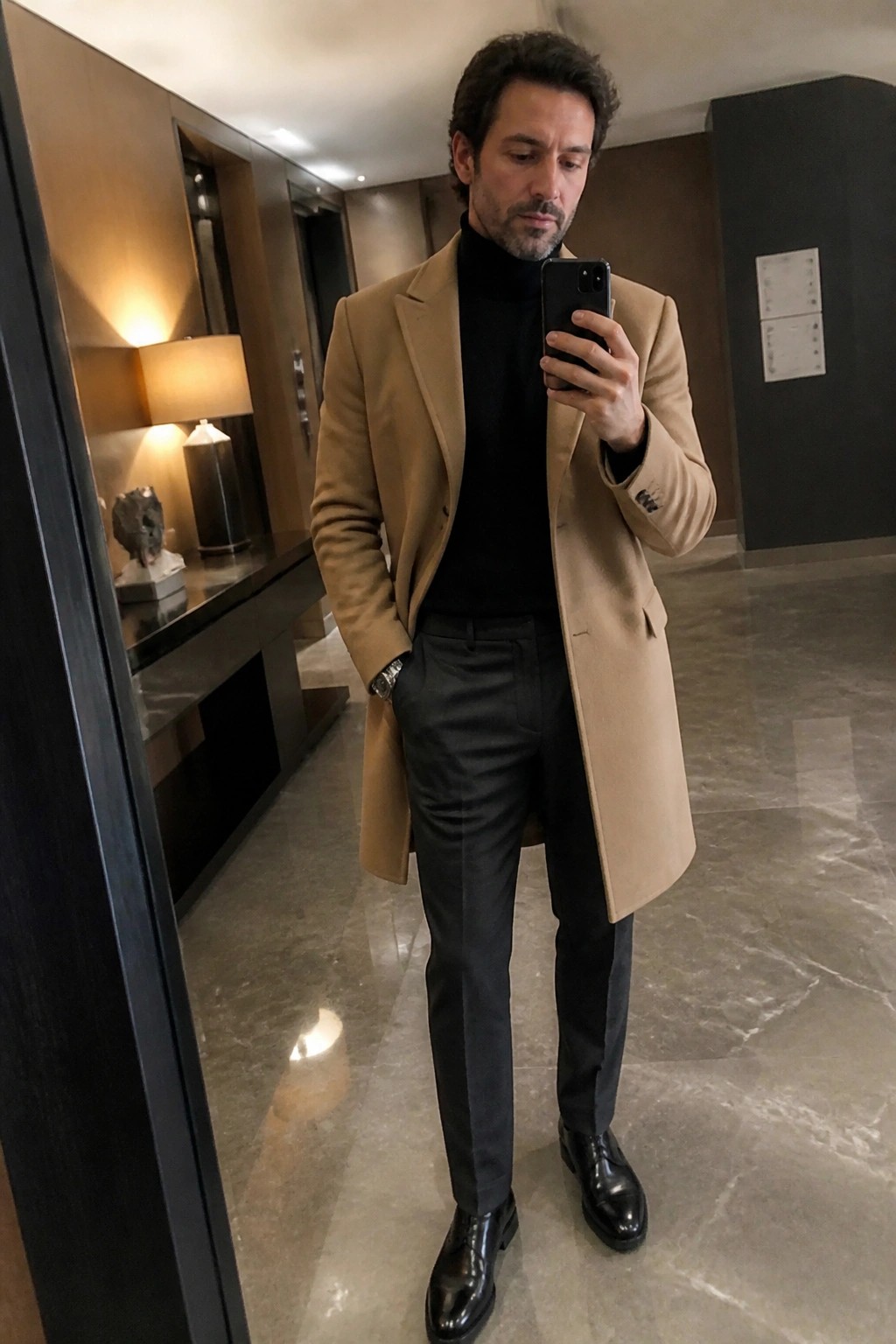 A man in a camel overcoat, black turtleneck, charcoal pants and black dress shoes holds a phone for a mirror selfie inside a modern lobby.
