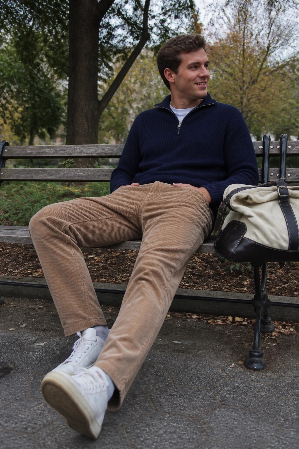 Man in navy quarter-zip sweater and tan corduroy pants on park bench with duffel bag.