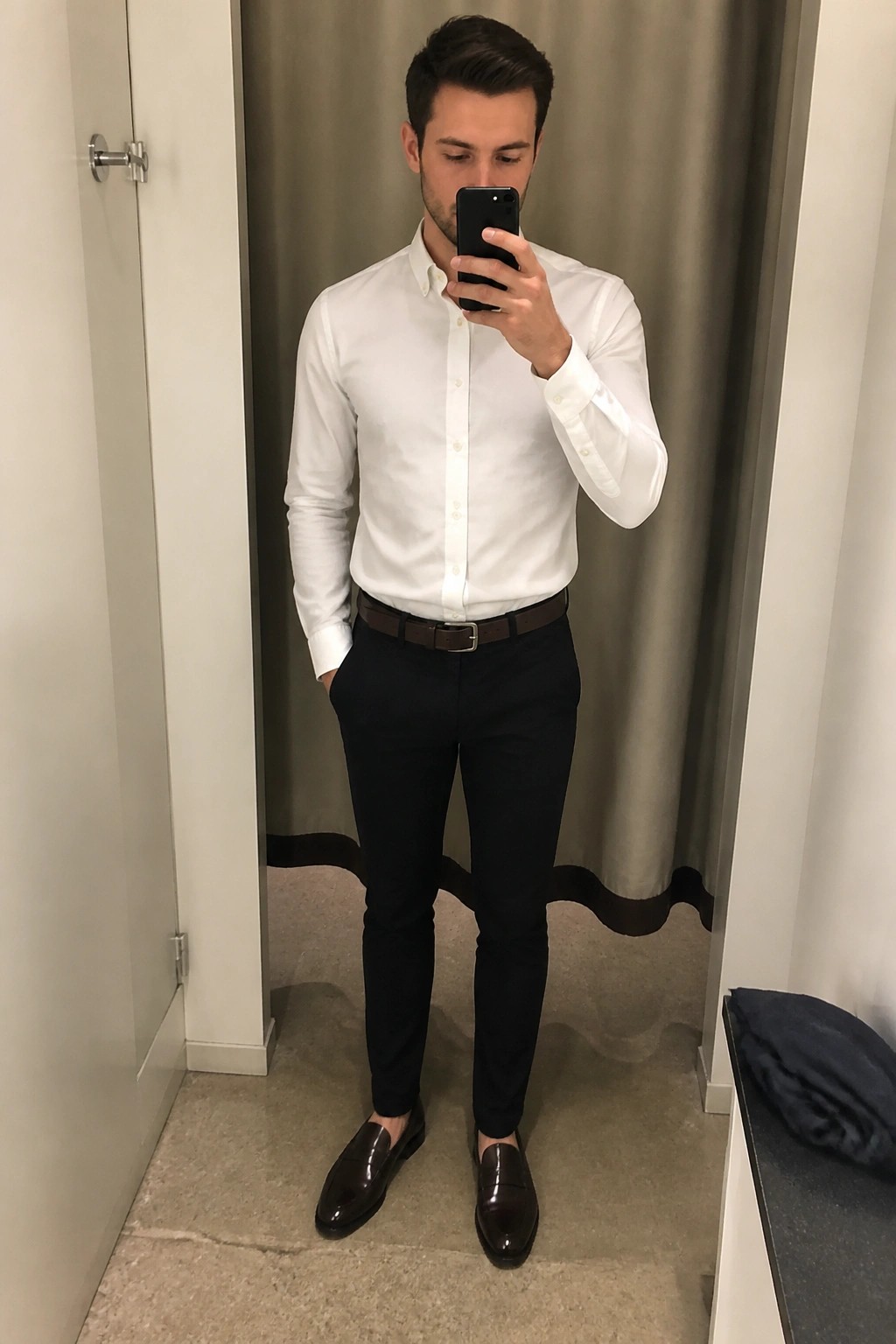 Man wearing slim black pants, white button-down shirt, brown leather belt, and brown loafers while taking a mirror selfie in a fitting room.