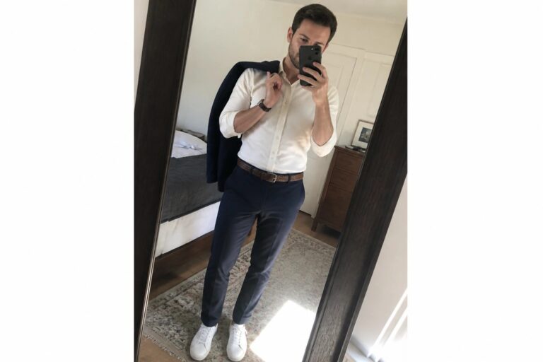 Man in beige shirt and navy pants taking mirror selfie while holding jacket