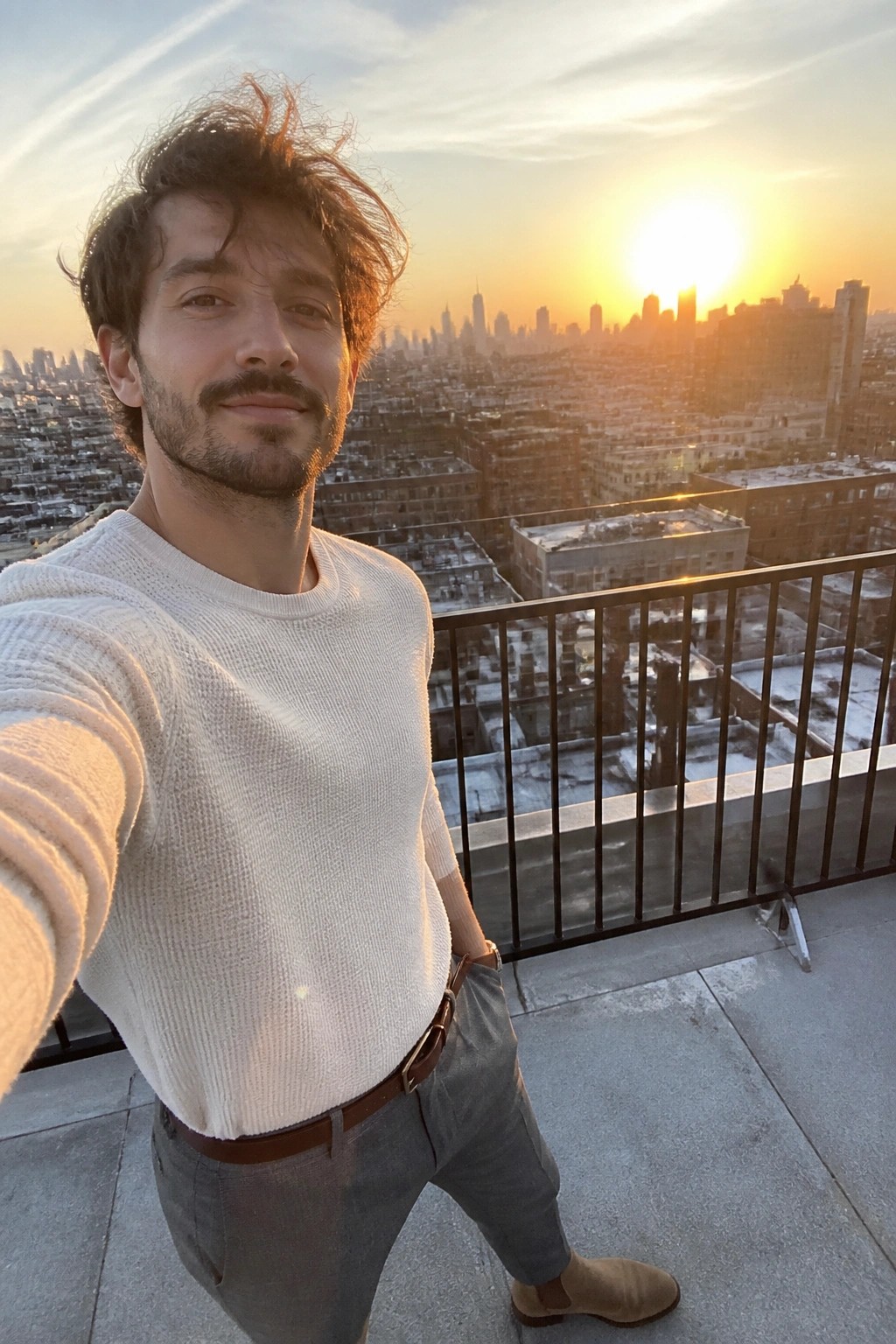 Man in white sweater posing on rooftop balcony at sunset over city skyline