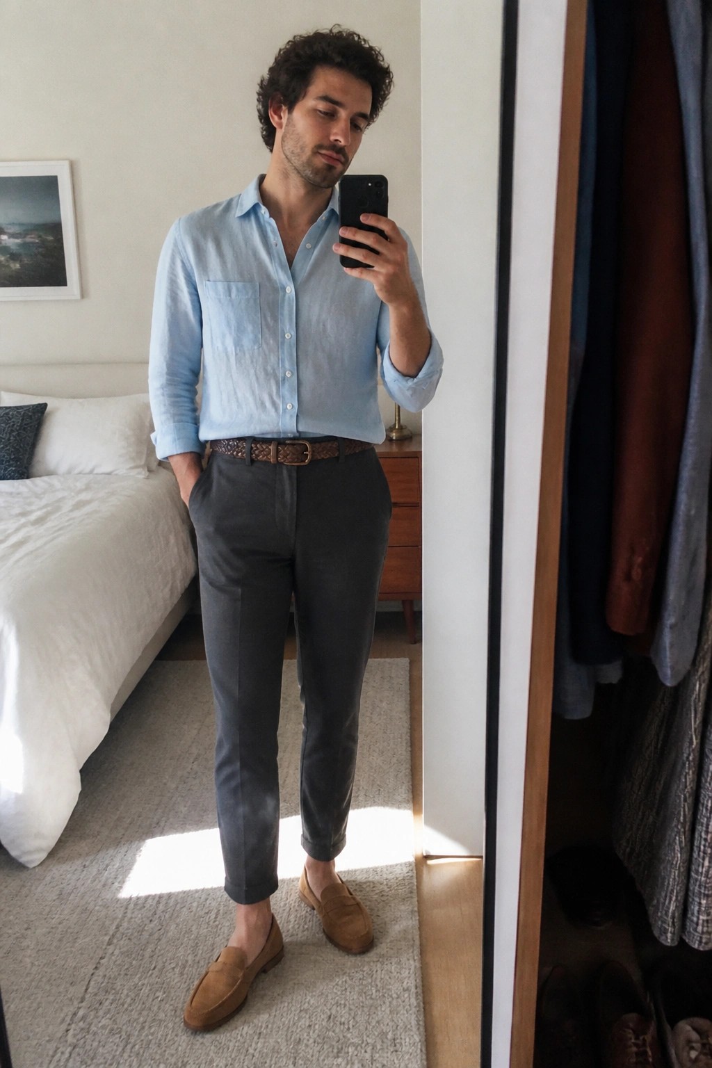 Man in light blue shirt and gray pants taking mirror selfie in bedroom