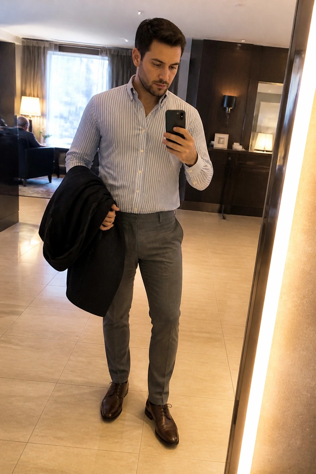 Man in striped shirt and gray pants holds black jacket while viewing phone in mirror
