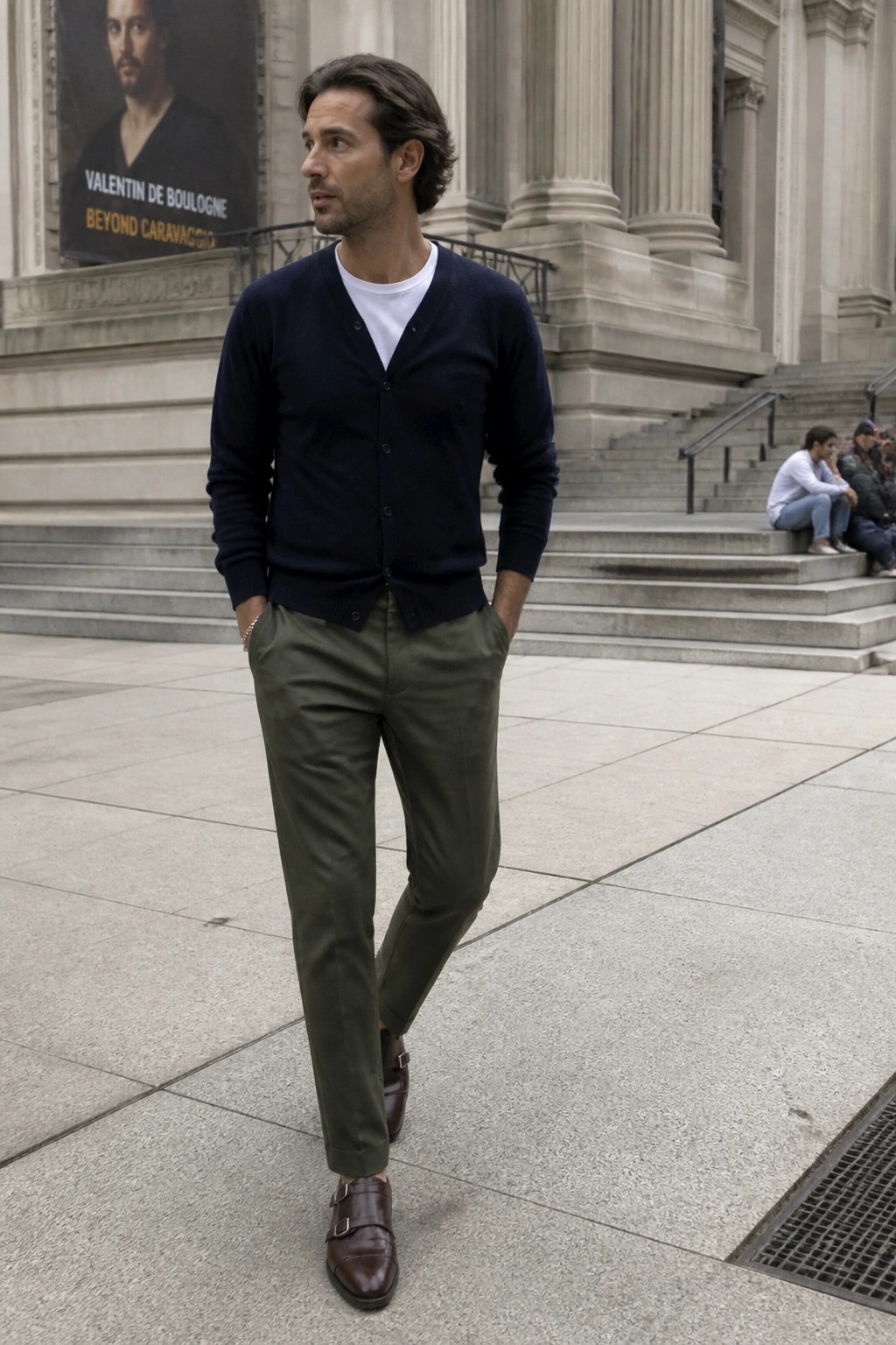 Man in navy cardigan, olive pants, and brown monk shoes before stone building.
