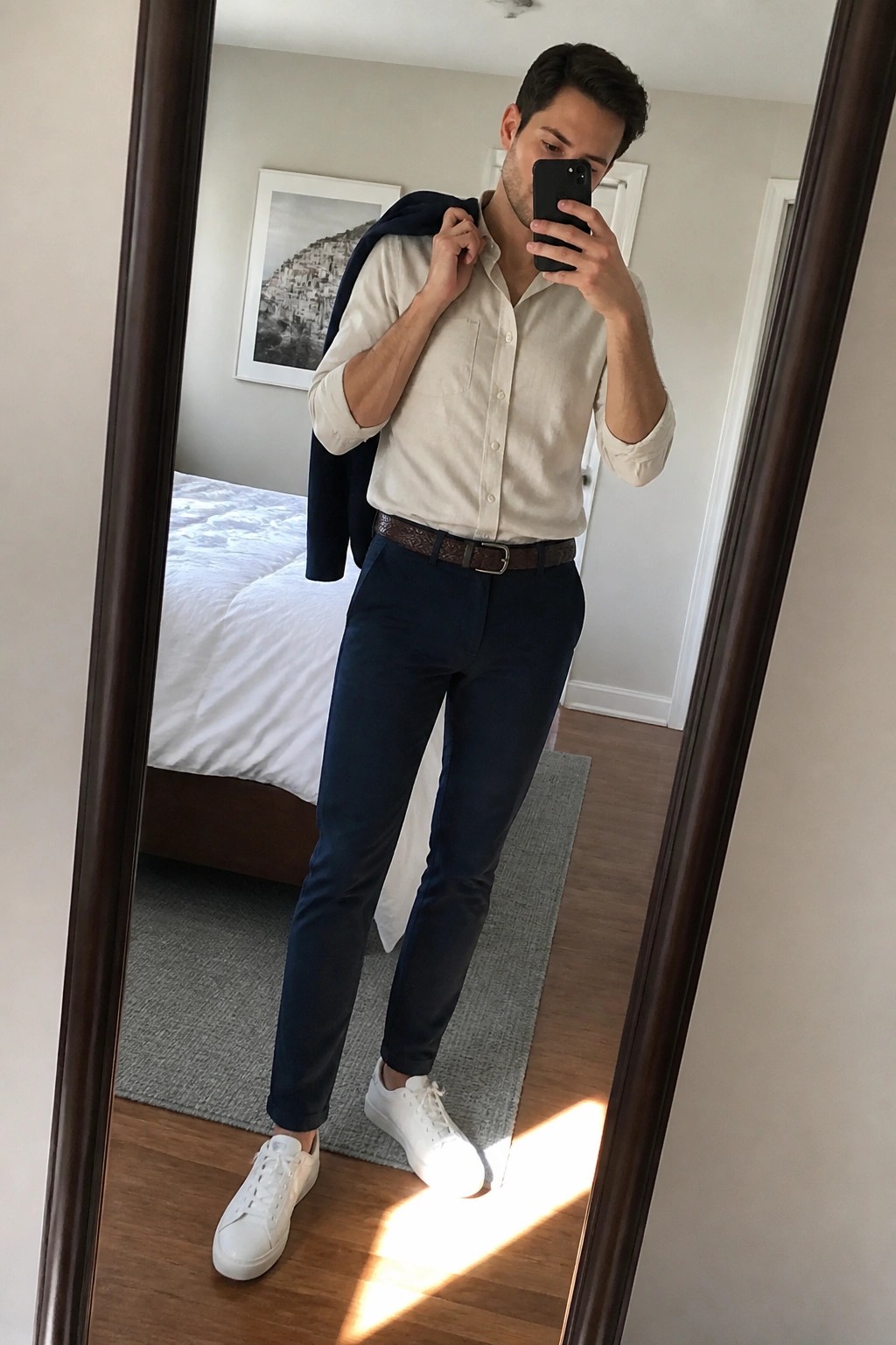 Man in beige shirt and navy pants taking mirror selfie while holding jacket