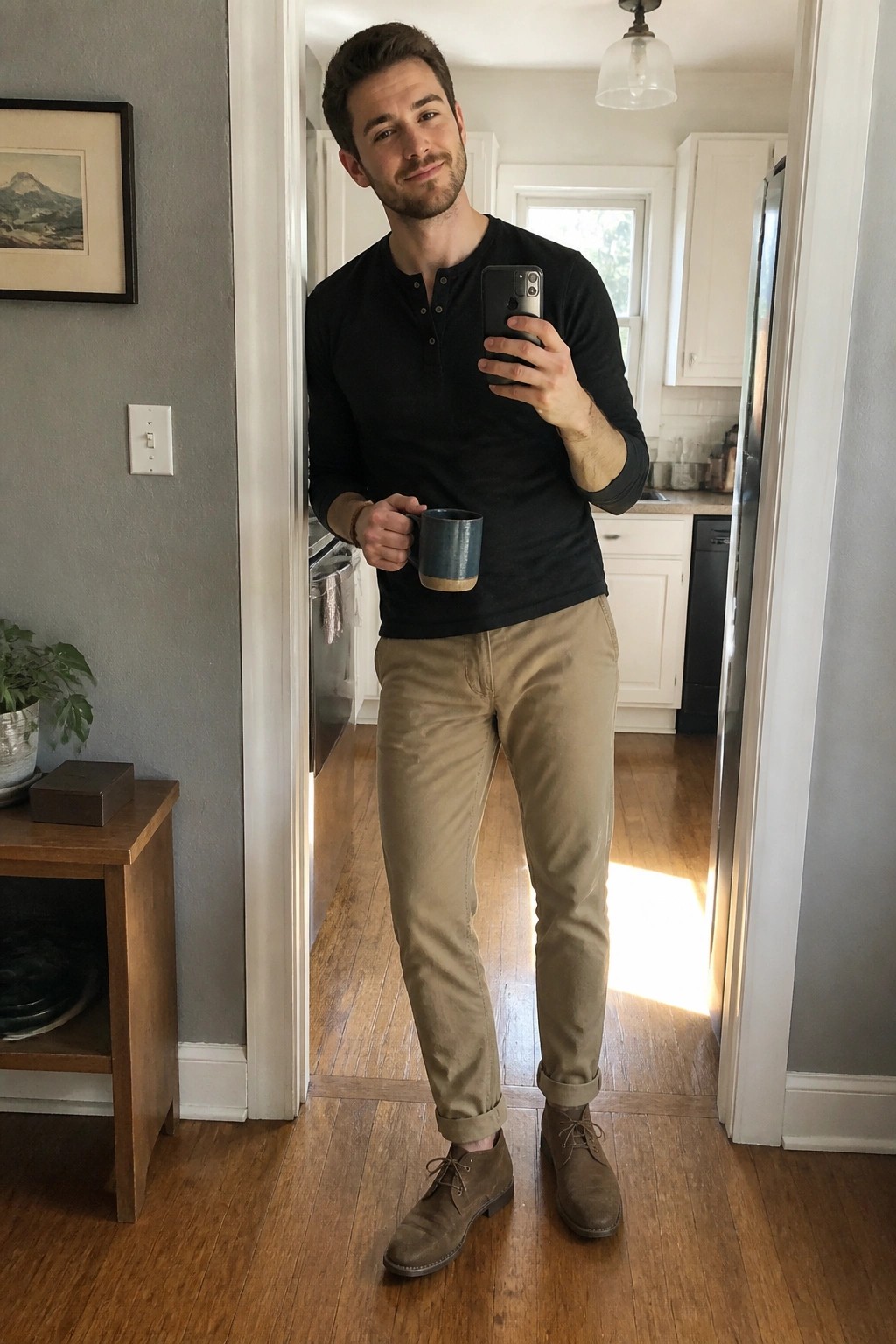 Man in black henley and khaki pants takes mirror selfie holding a mug