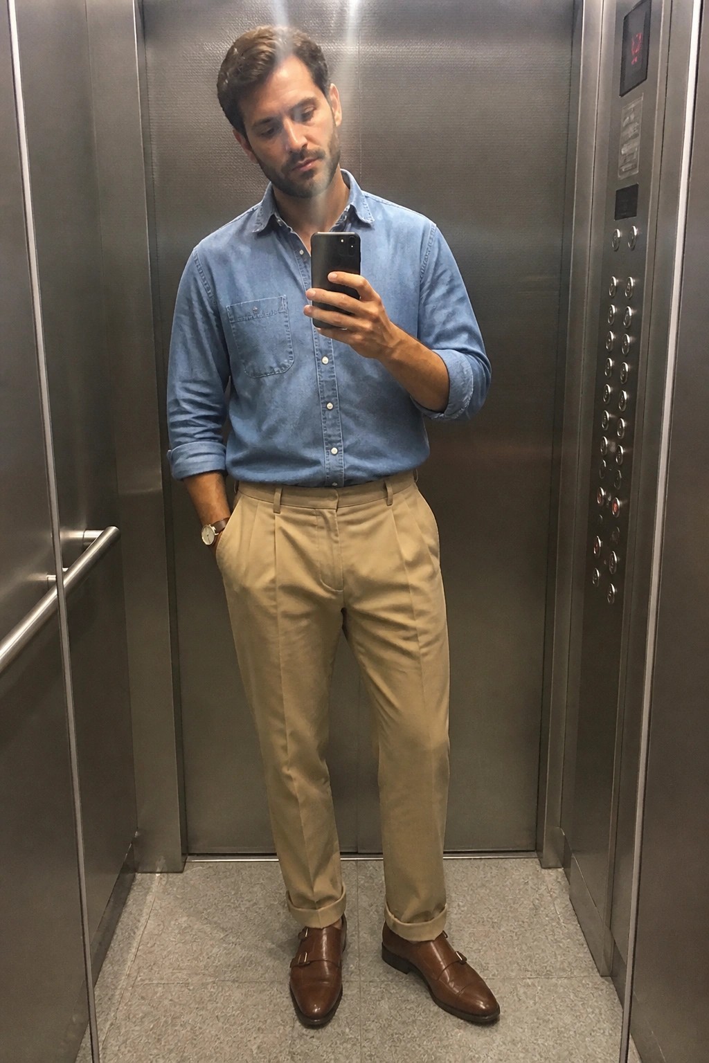 man standing in elevator wearing light blue chambray shirt with rolled sleeves, beige pleated relaxed fit pants cuffed at hem, brown leather double monk strap shoes, and wristwatch while holding phone