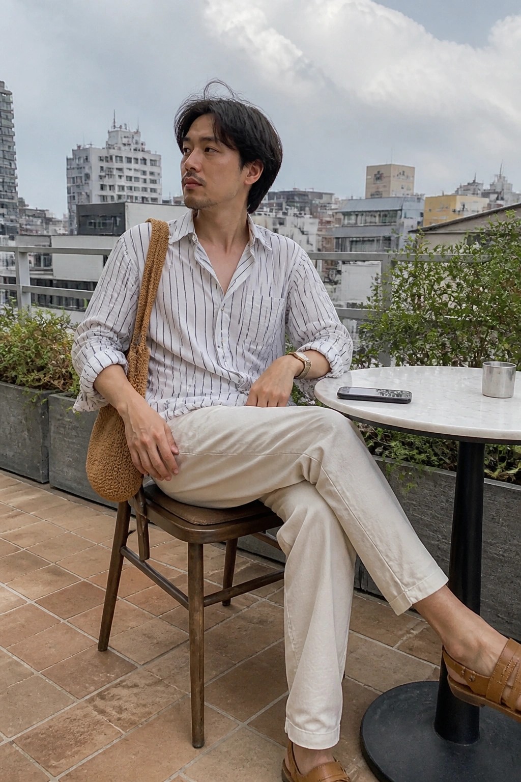 Man in beige relaxed fit pants and white striped shirt seated with brown shoulder bag and sandals visible.