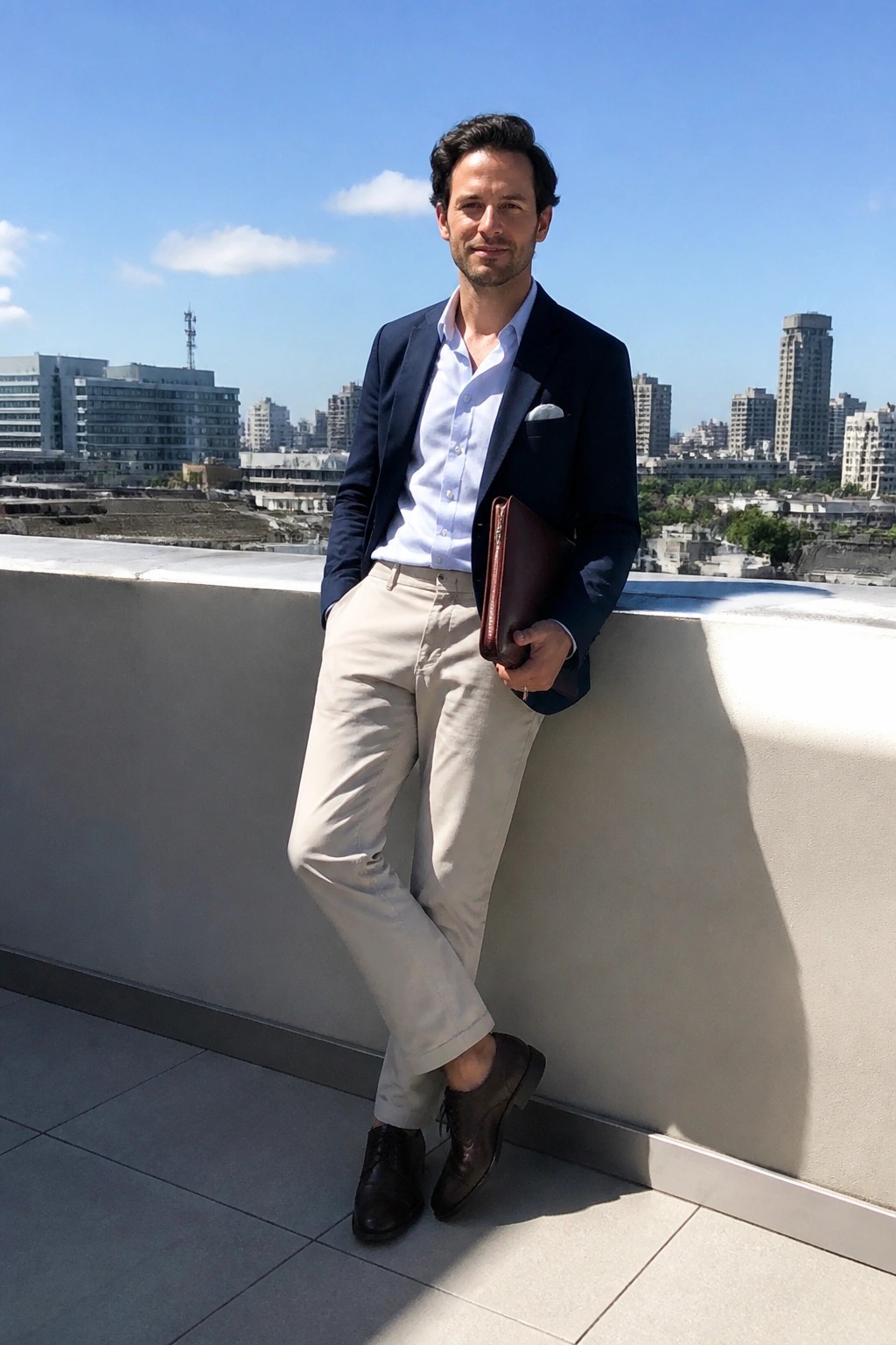 Man in navy blazer and beige pants holding leather portfolio on sunny city balcony