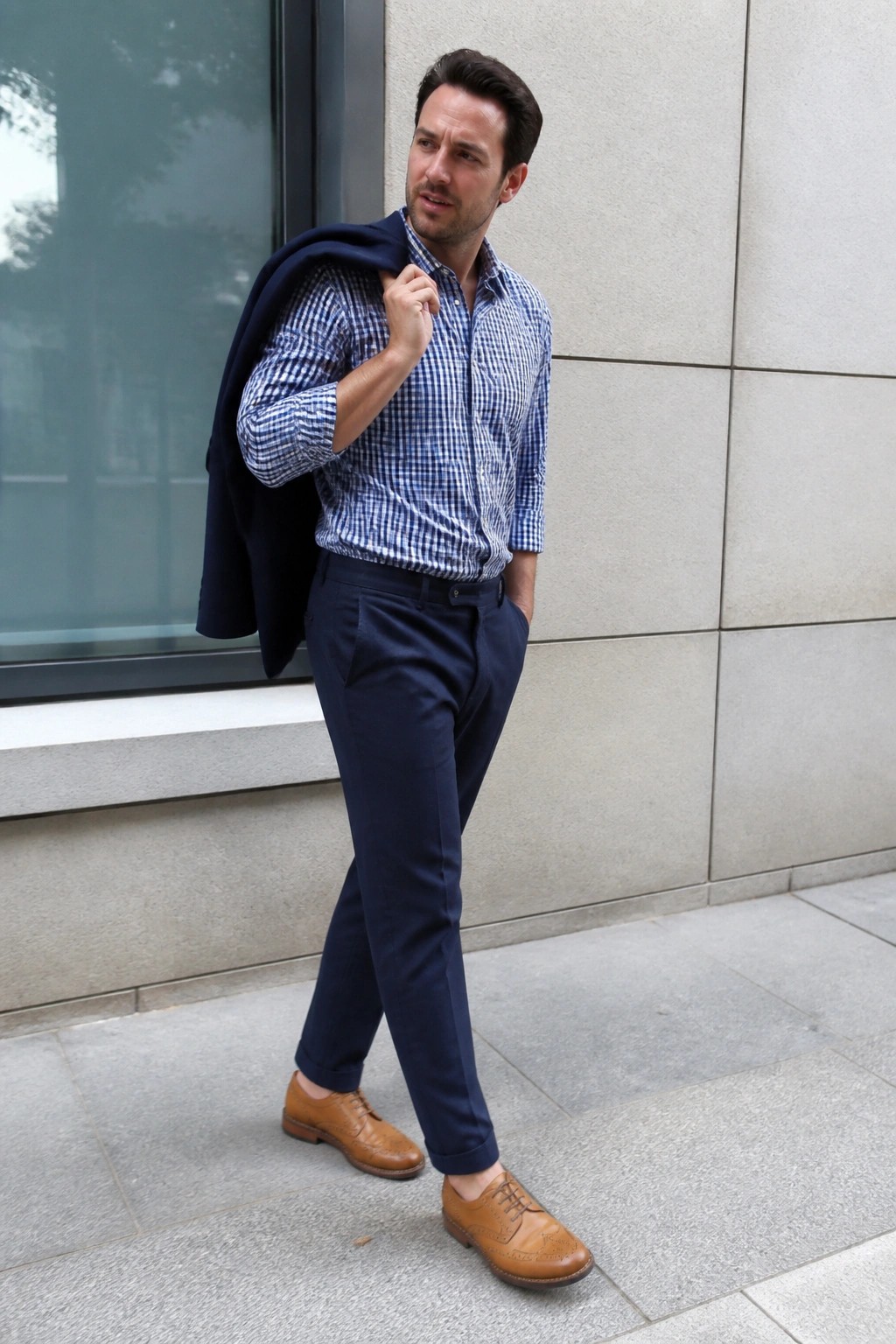 Man in navy relaxed fit formal pants, blue white gingham shirt with rolled sleeves, brown brogue shoes, and navy blazer held over shoulder in office appropriate smart casual style