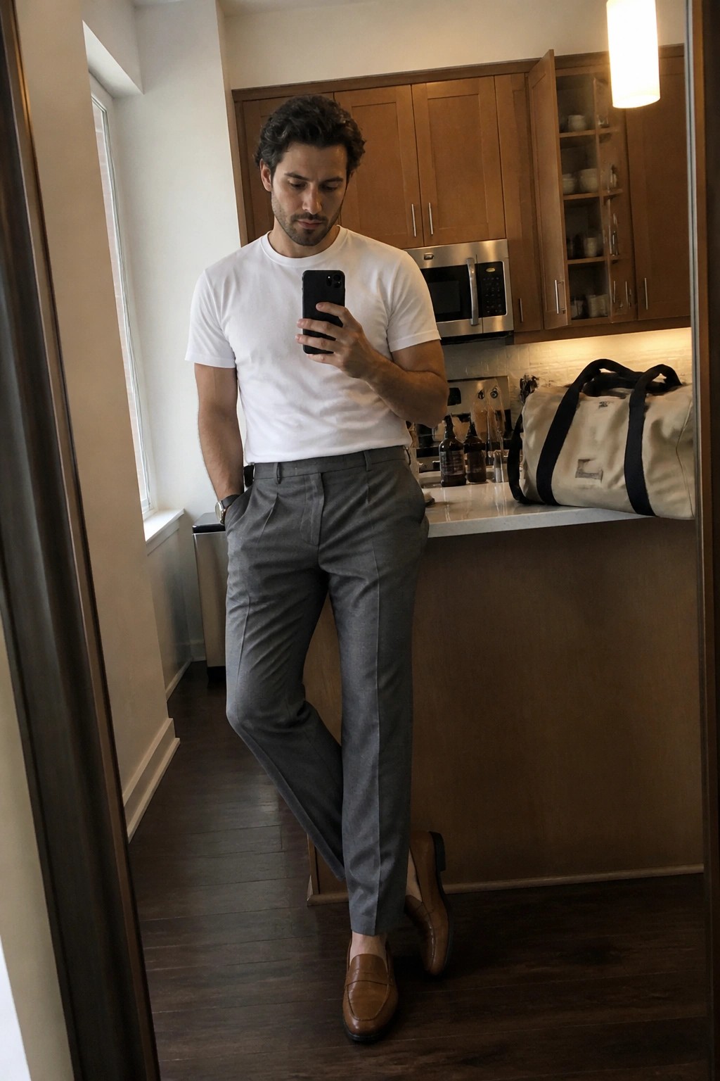 Man wearing relaxed fit gray pleated trousers, white crewneck t-shirt, and brown leather loafers while taking a mirror selfie in a kitchen setting.