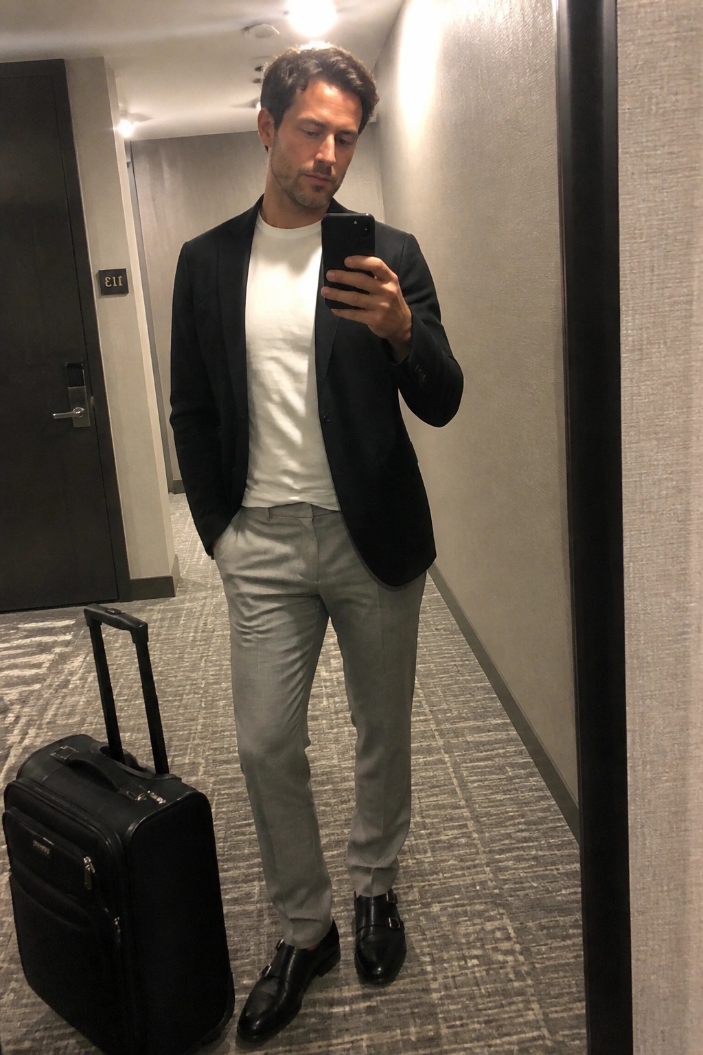 Man in black blazer, white tee, gray pants with suitcase in hallway.