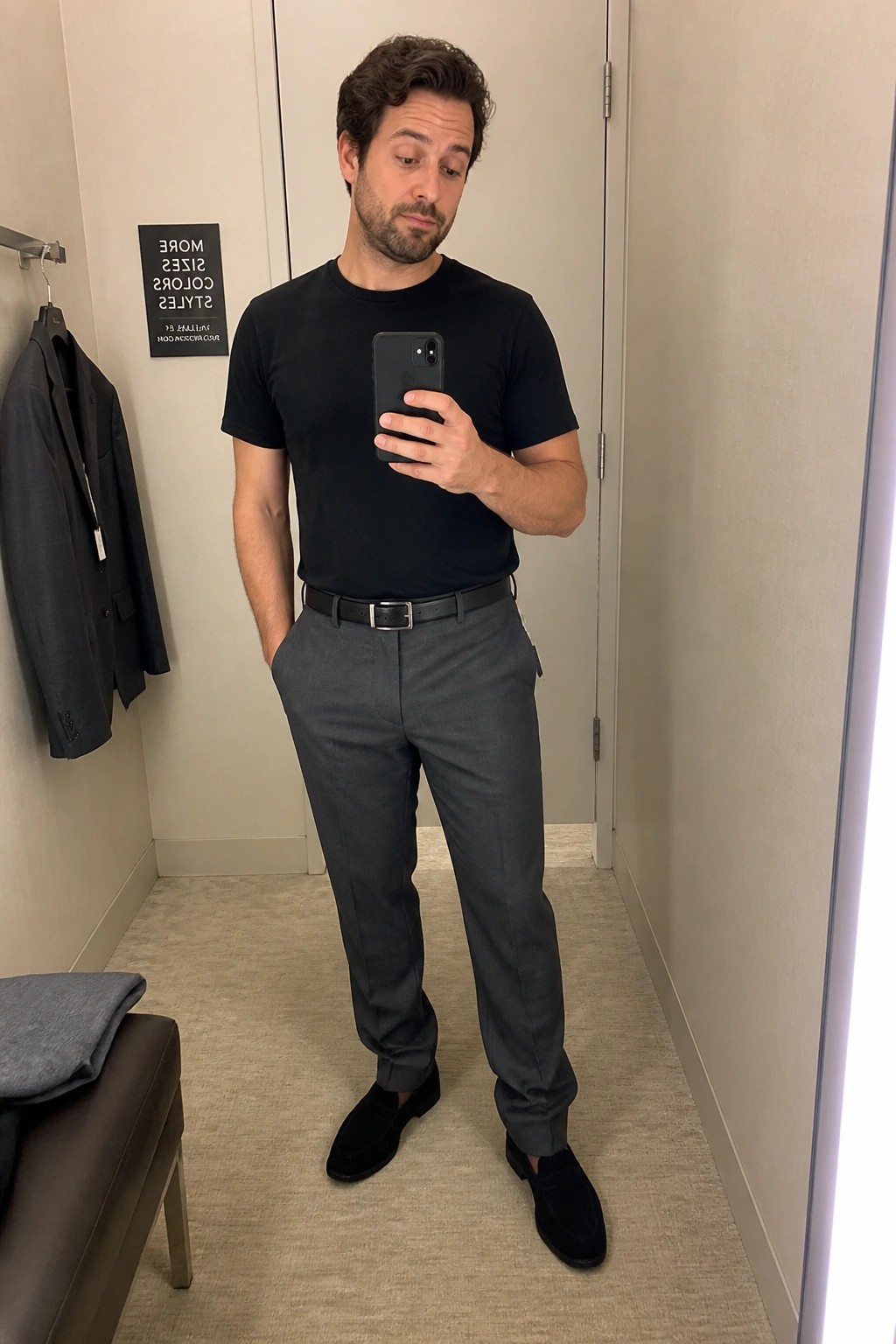 Man in black t-shirt and gray pants taking mirror selfie in fitting room