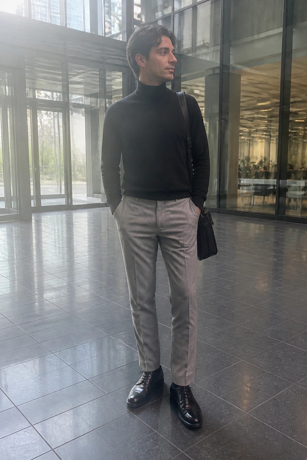 Man in black turtleneck and gray trousers with bag in glass atrium