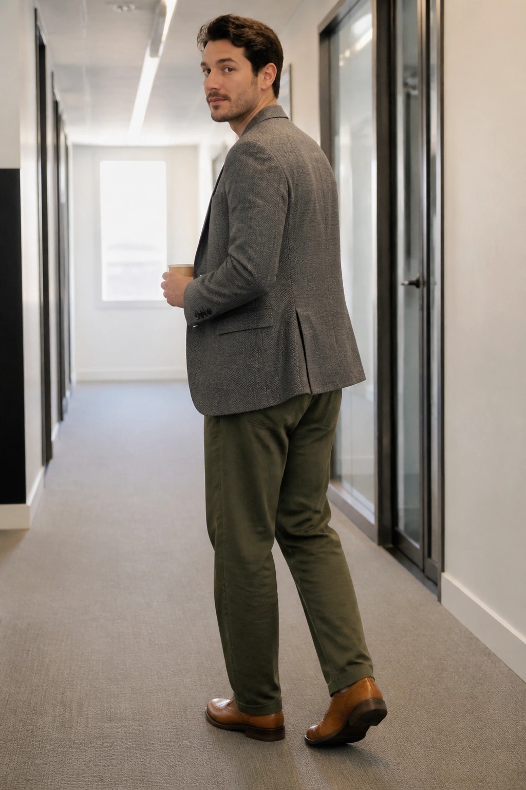 Man in gray blazer and green pants holds coffee glancing over shoulder in hallway