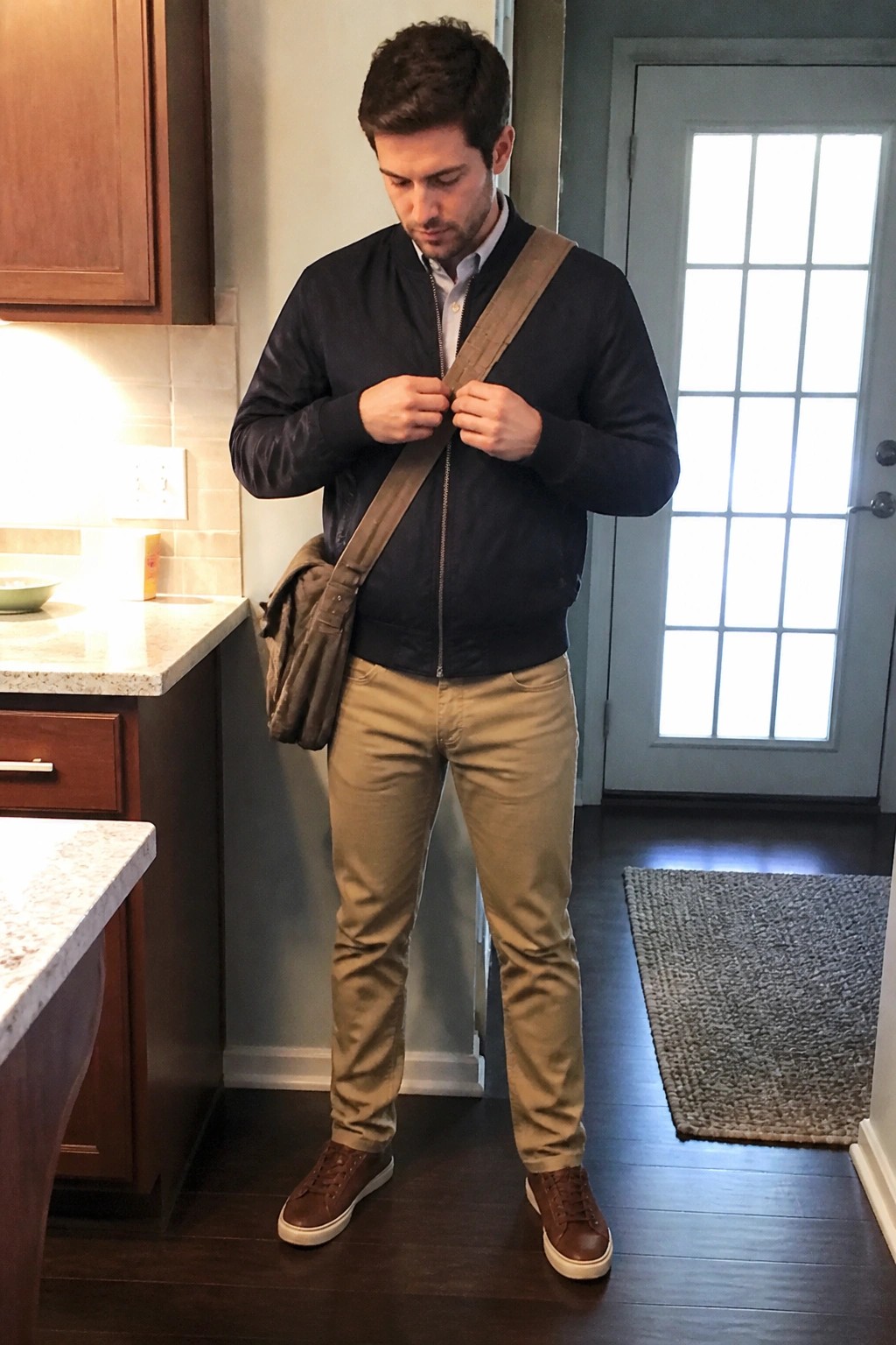 Man in navy bomber jacket and khaki pants adjusting brown messenger bag strap