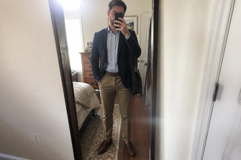 Man in navy blazer and khaki pants taking mirror selfie in bedroom