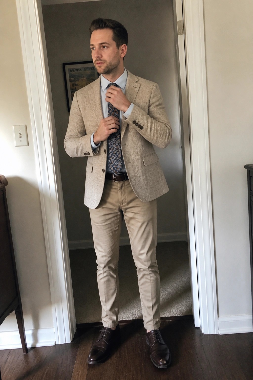 Man in beige blazer and khakis adjusts patterned tie in doorway