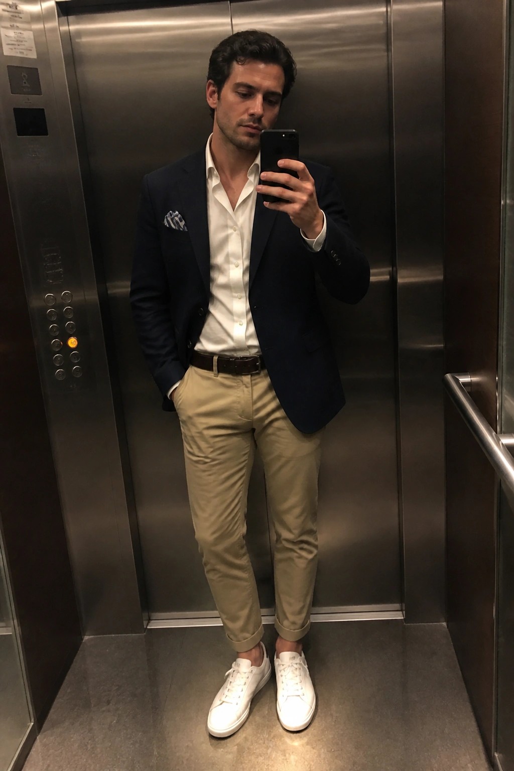 Man in navy blazer, open white dress shirt, khaki pants, brown belt, and white sneakers holding a phone for a mirror selfie inside an elevator.