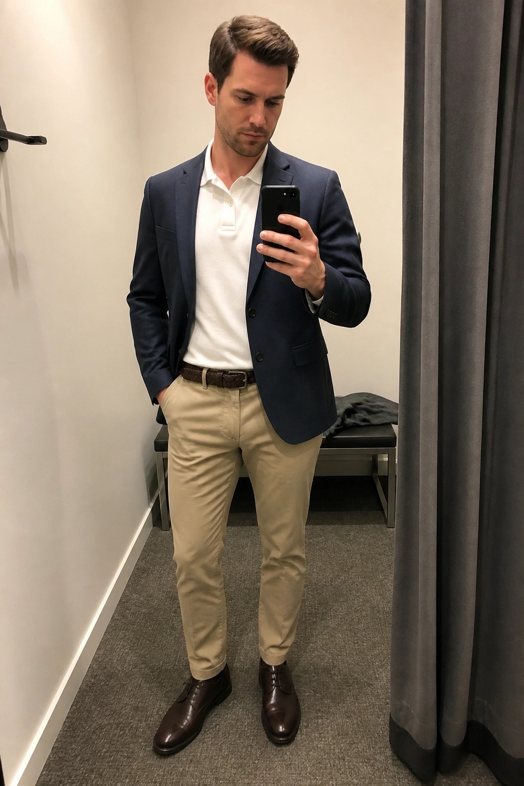 Man in navy blazer over white polo shirt with khaki pants brown belt and brown dress shoes taking a mirror selfie in a fitting room