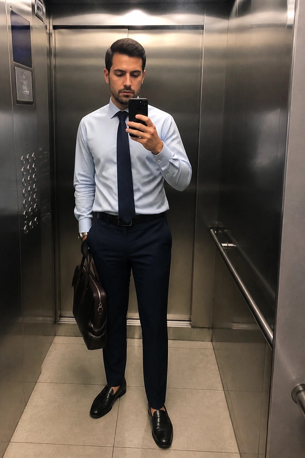 Man in light blue shirt and navy tie takes selfie in elevator with bag.