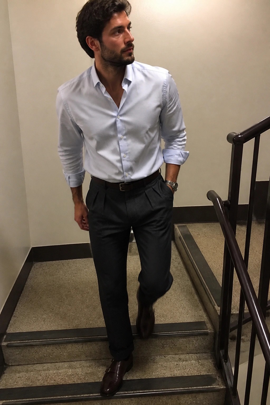 Bearded man in light blue shirt and dark pleated pants on staircase.