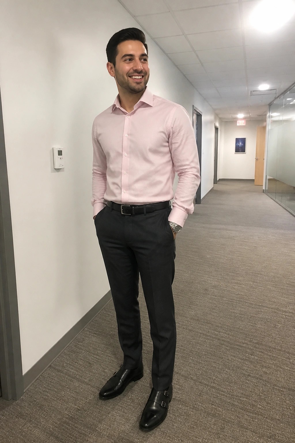 Smiling man in pink shirt and black trousers stands in office hallway.