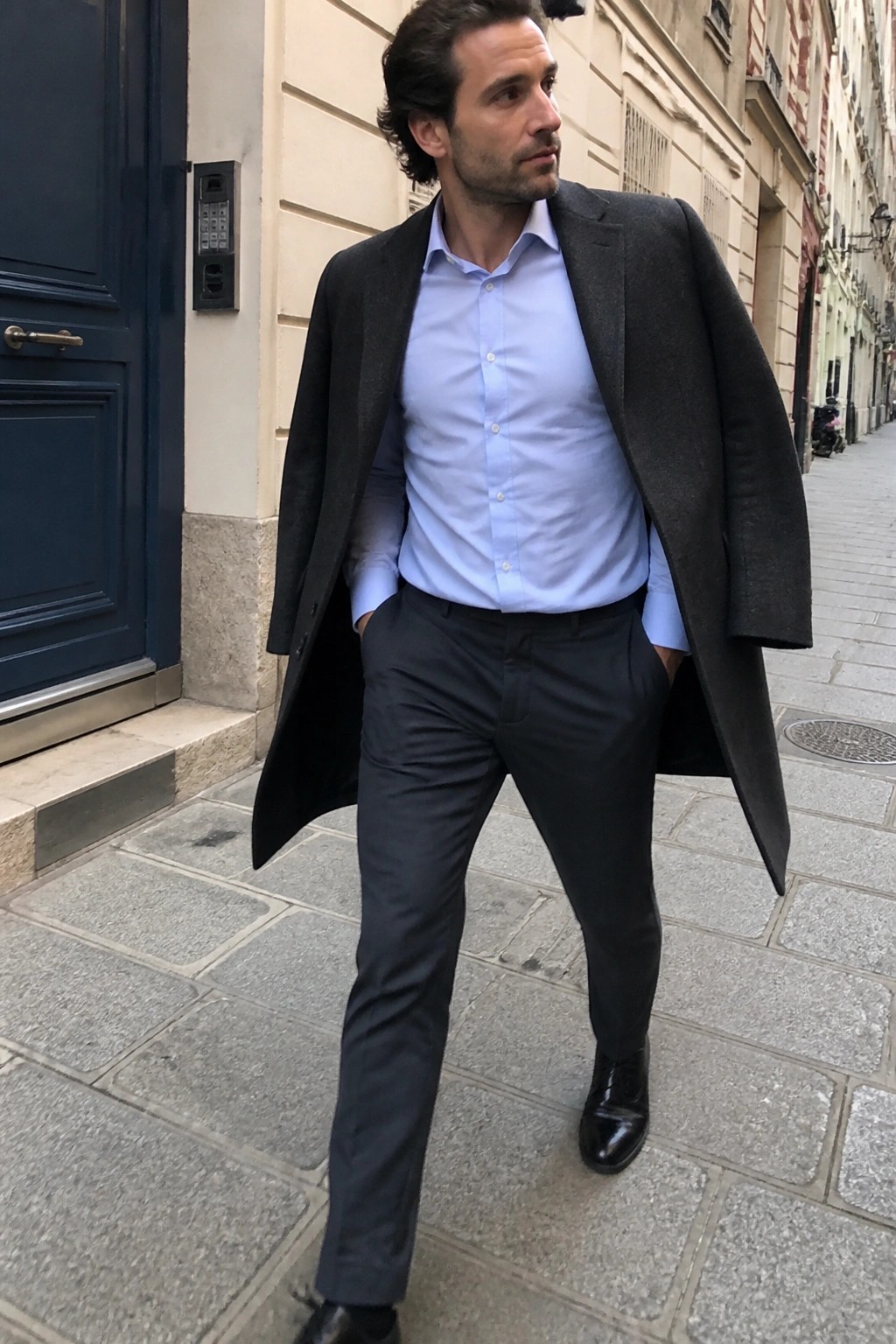 Man in light blue shirt and open dark overcoat on cobblestone street with hands in pockets