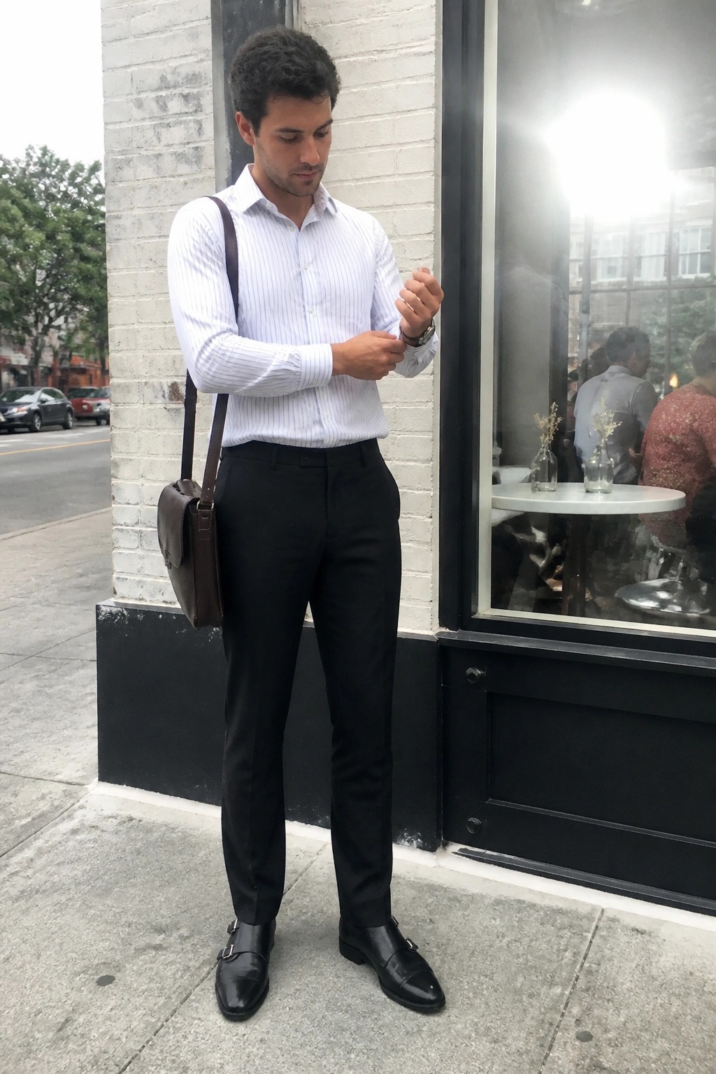 Man in white pinstriped button down shirt tucked into black trousers with black double monk strap shoes and brown leather messenger bag over one shoulder standing on sidewalk.